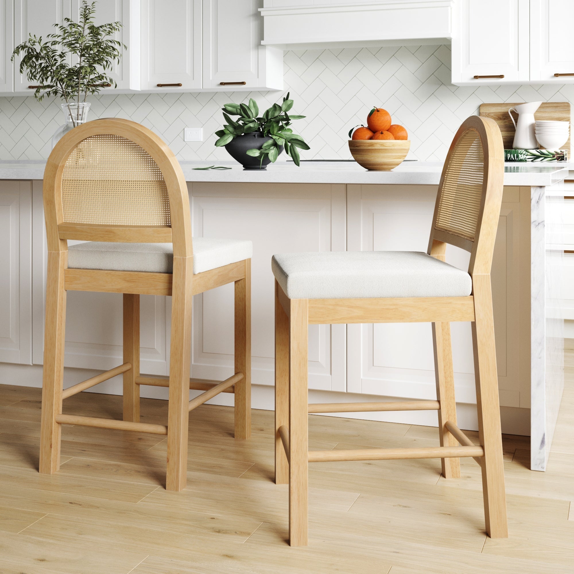 Two Nathan James Boucle & Rattan Counter Height Bar Stools in Warm Pine are placed by a white kitchen island with white cabinets, herringbone tile backsplash, and bowls of fruit and greenery on the counter.