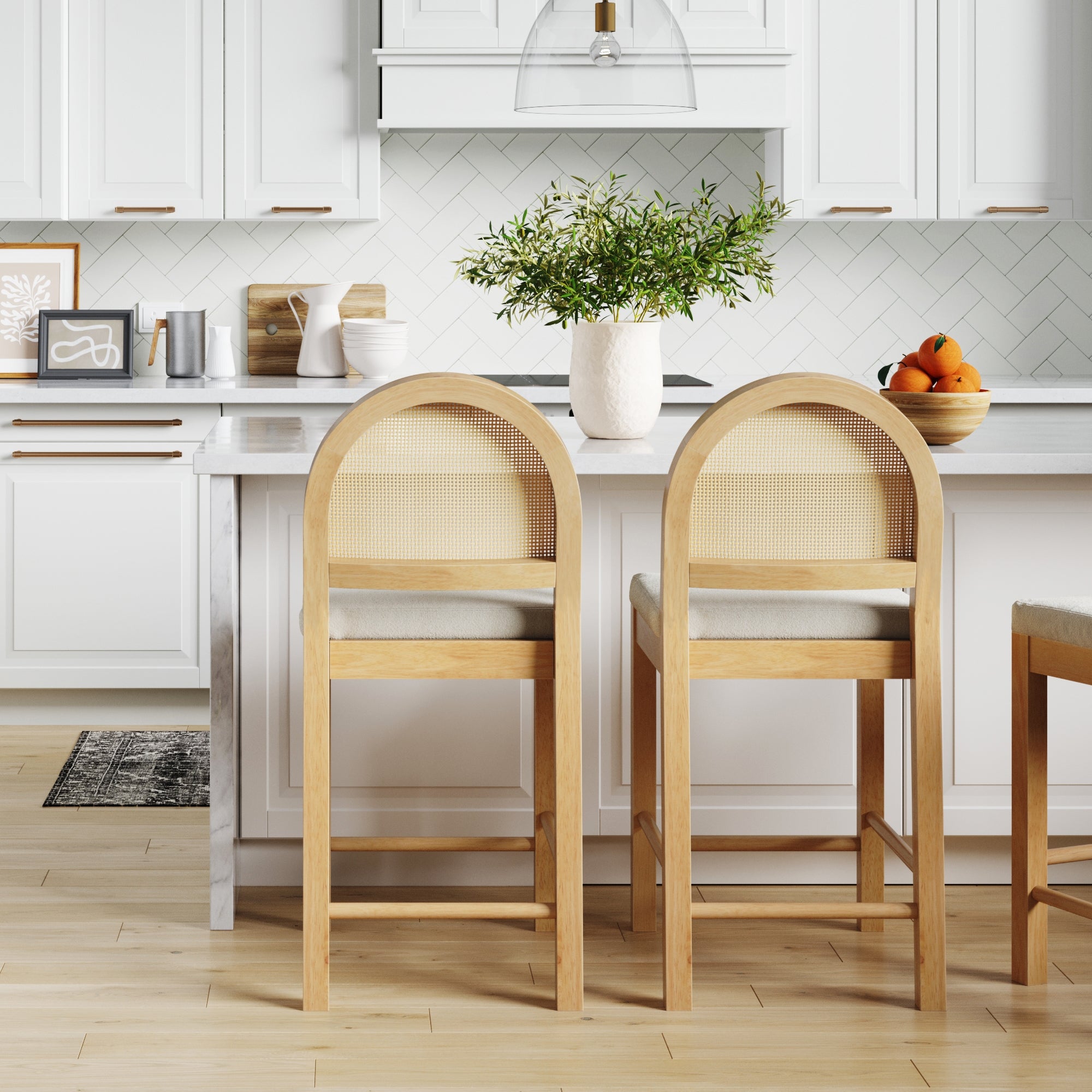 A modern kitchen with white cabinets and a tile backsplash features two Nathan James Boucle & Rattan Counter Height Bar Stools in Warm Pine at a marble island, topped with a white vase of green branches and a bowl of oranges.