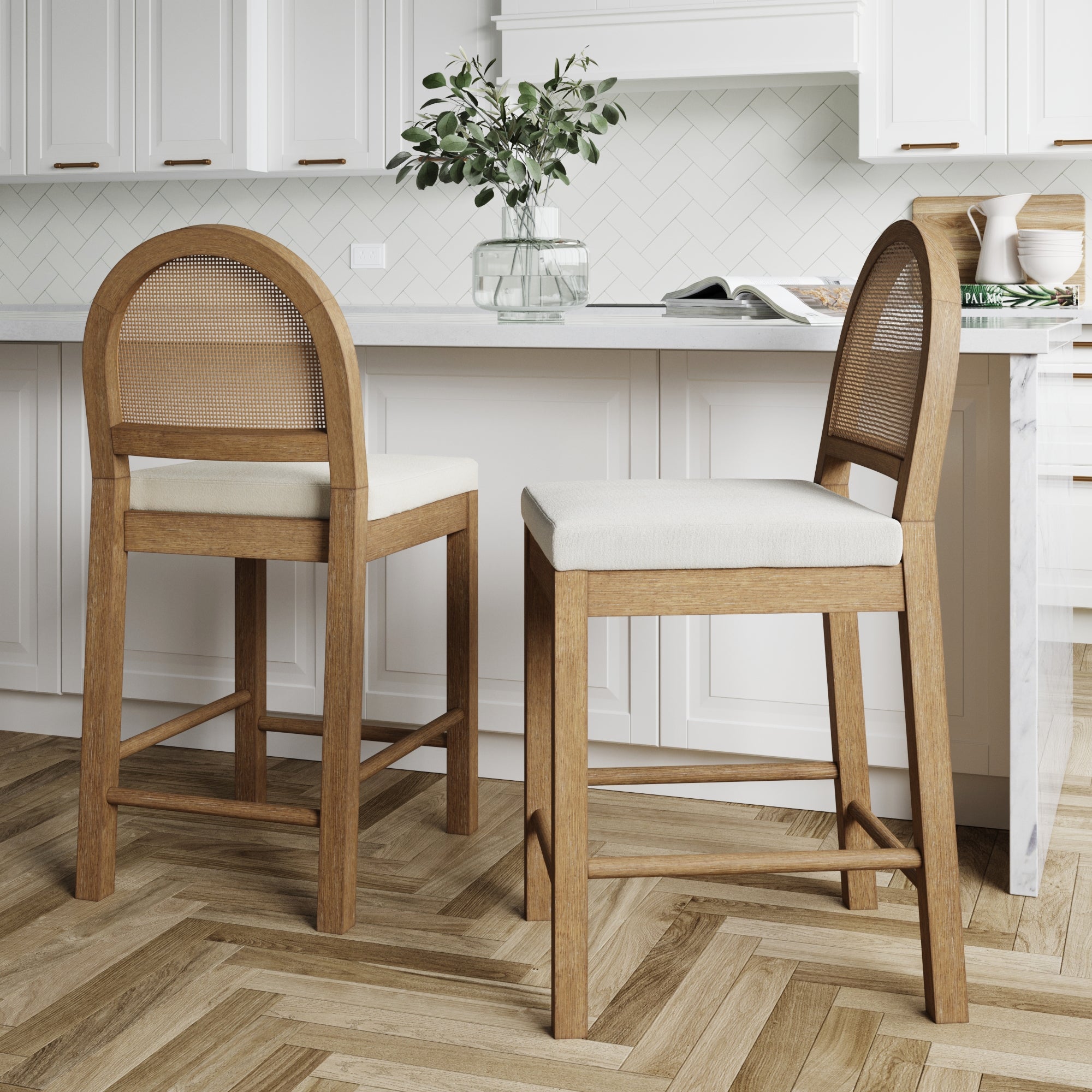 Two wooden bar stools with curved cane backs and cushioned seats are placed by a white kitchen island. The kitchen features white cabinets, herringbone wood flooring, and a glass vase with greenery on the counter.