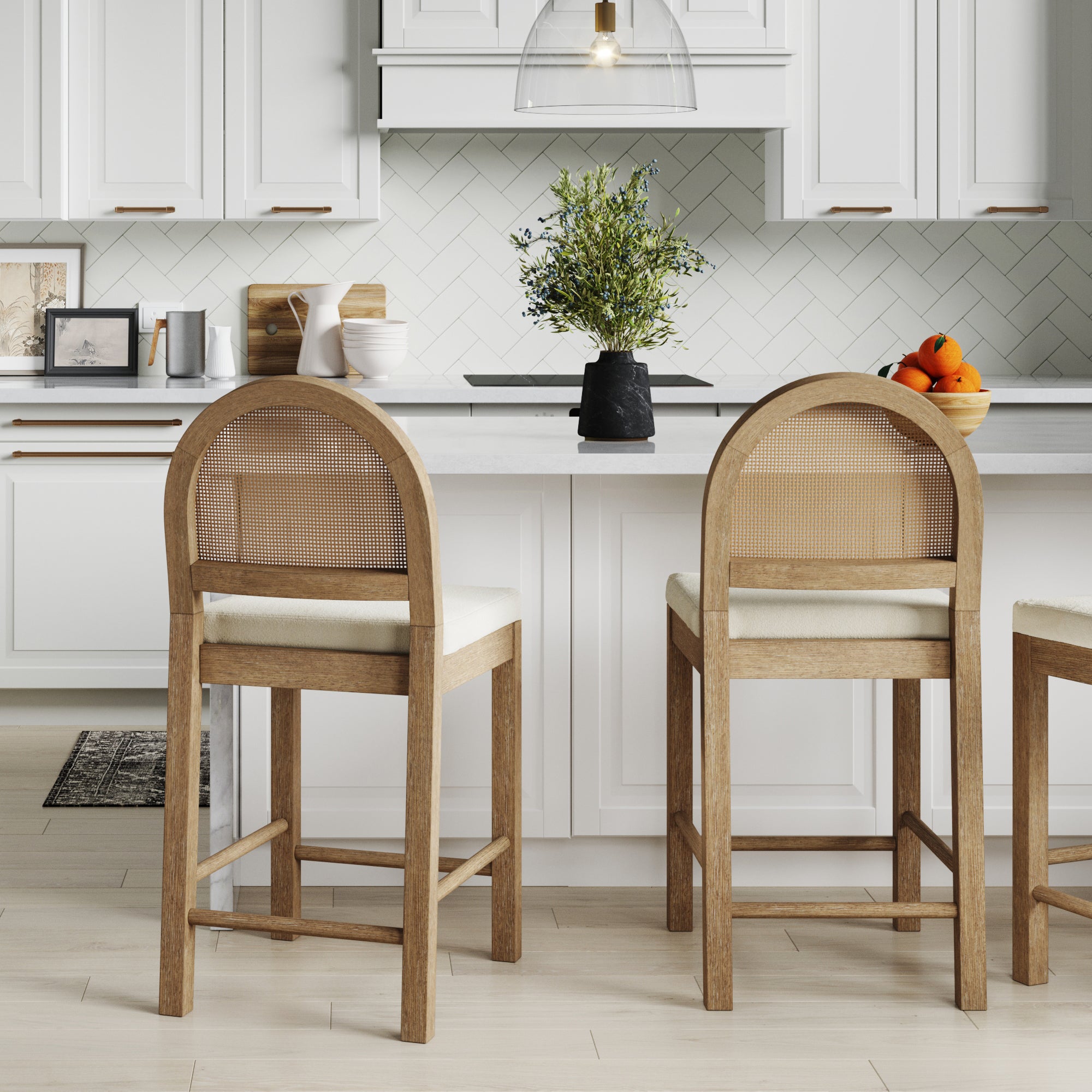 Two Boucle & Rattan Counter Height Bar Stools by Nathan James sit at a white kitchen island with white cabinets, a herringbone tile backsplash, and decor like a black vase with greenery and oranges for a bohemian touch.