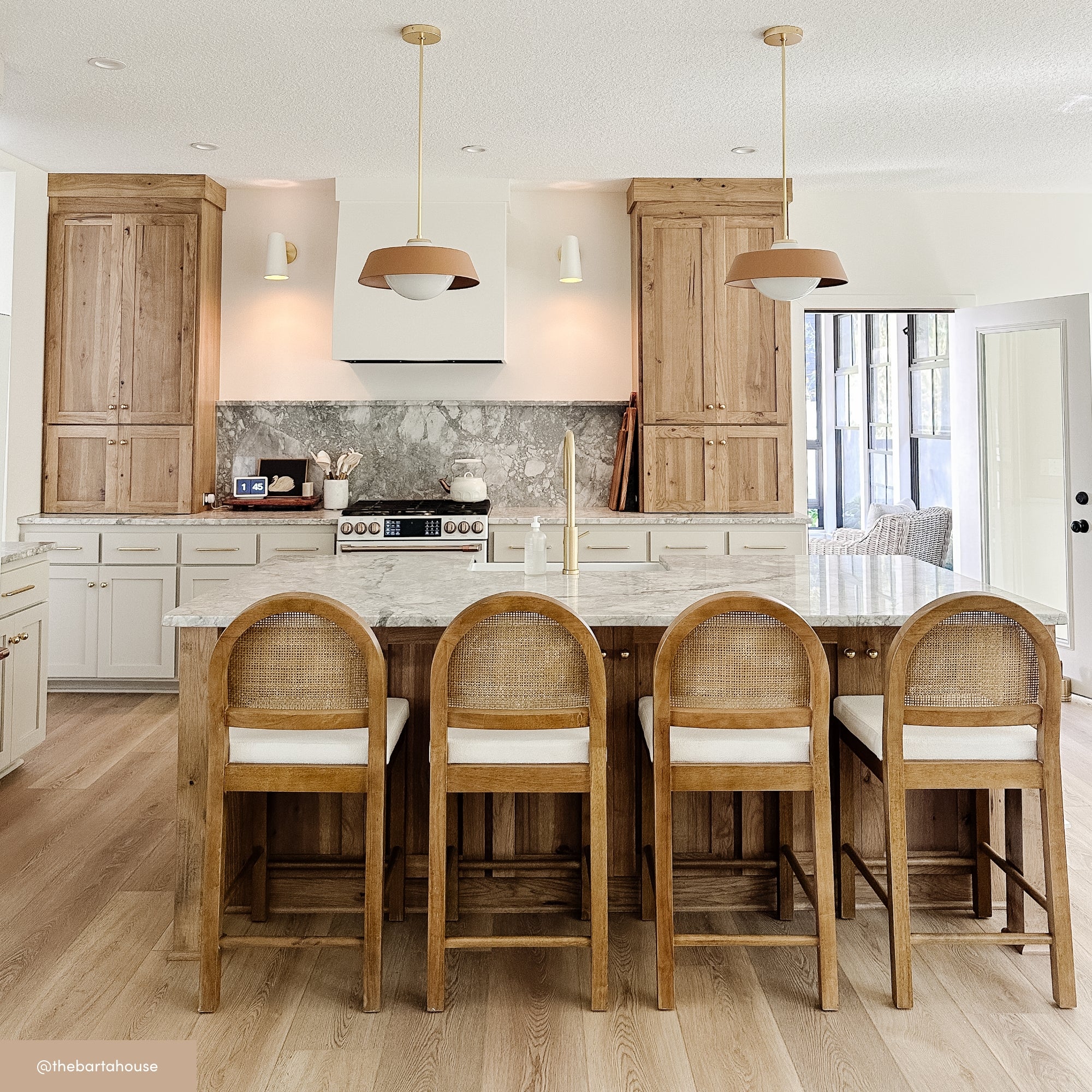 Bright, modern kitchen with light wood floors, a marble island, and four wooden barstools. Natural wood cabinets, white lower cabinets, pendant lights, and a marble backsplash create a warm, inviting space.