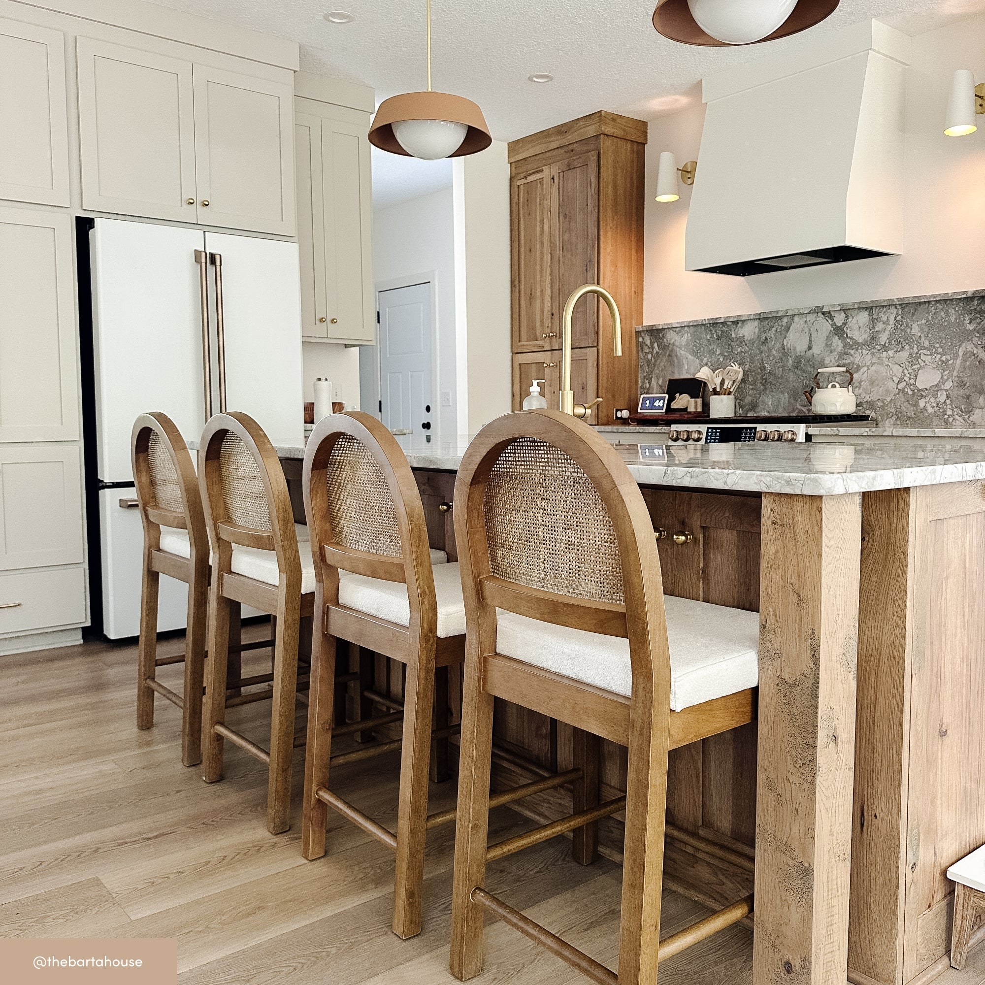A modern kitchen with light wood floors, an island with four wooden barstools, white cabinetry, a white range hood, marble backsplash, gold faucet, and pendant lights. A refrigerator and built-in oven are visible.
