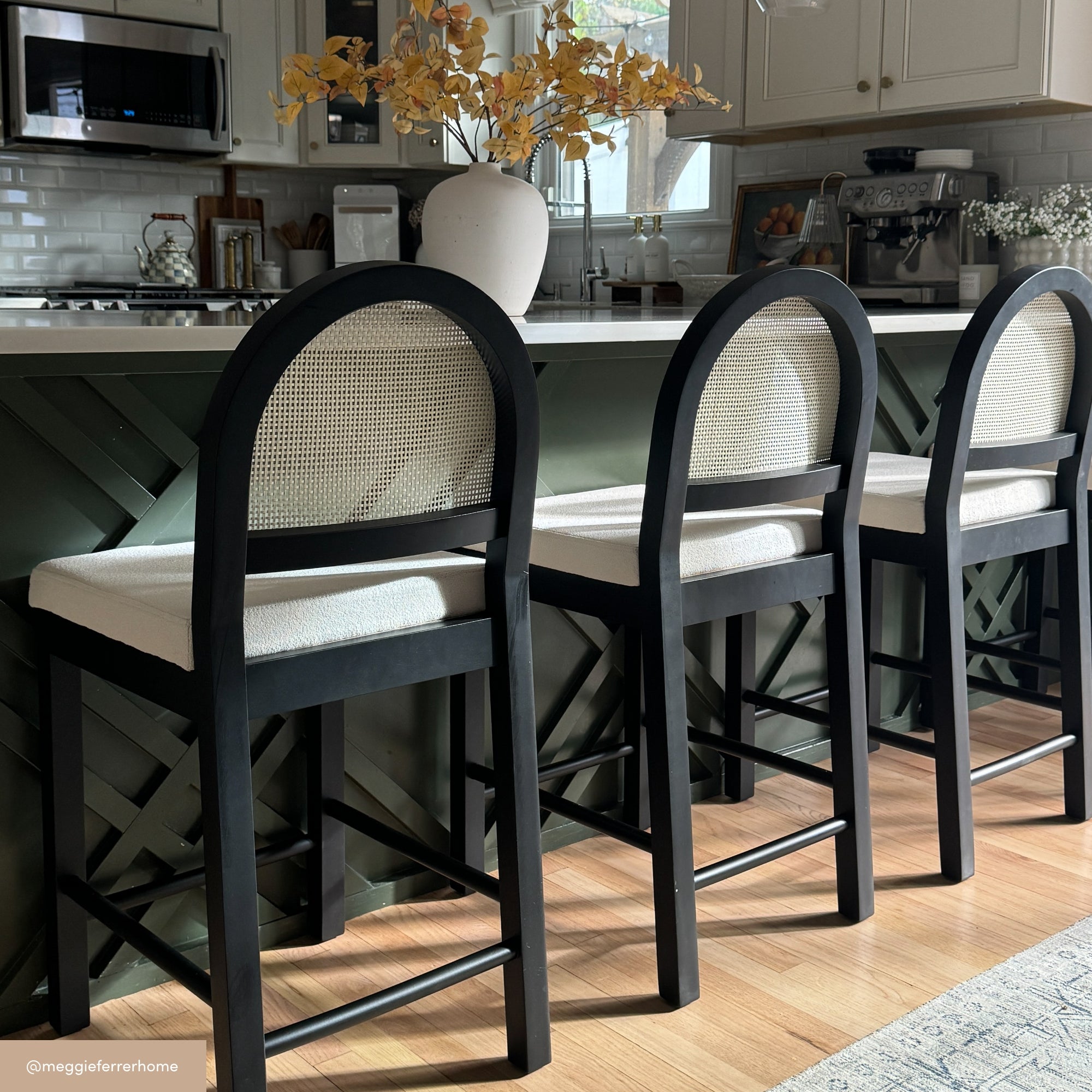 Three modern barstools with black frames and white upholstered backs and seats are lined up at a kitchen island with a green geometric-patterned front. A vase with autumn leaves sits on the countertop.