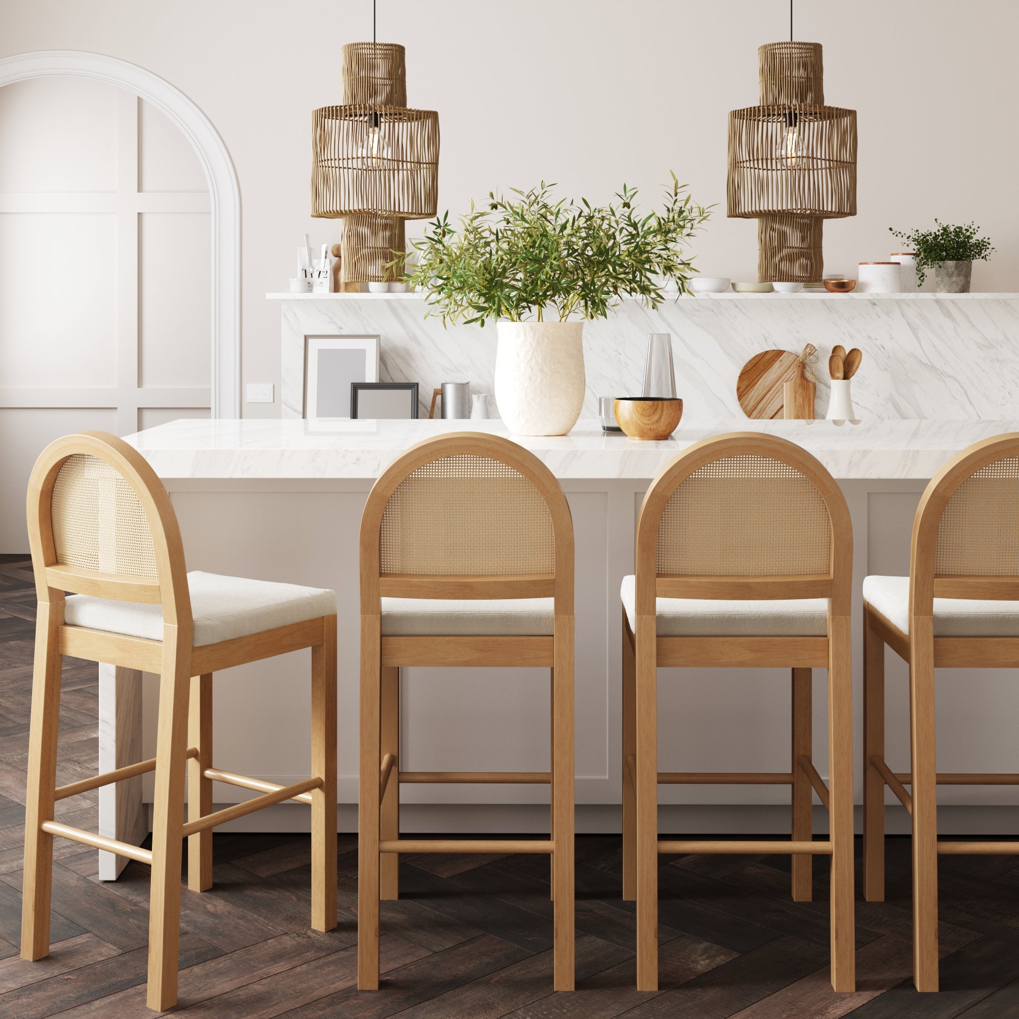A modern kitchen features a white marble island, four Nathan James Boucle & Rattan Bar Stools in Warm Pine, a large greenery-filled vase, and two woven pendant lights above—all in neutral tones with minimal decor.