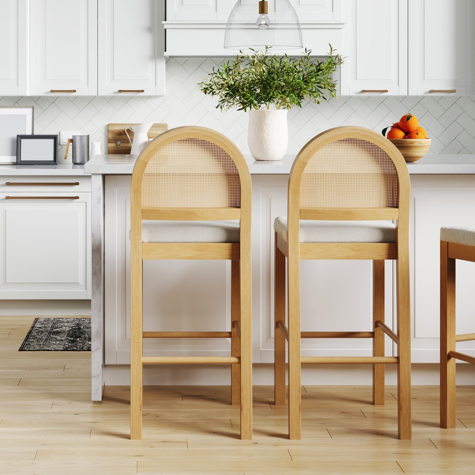 Bright kitchen with light wood floors, white cabinets, and a white countertop. Two Nathan James Boucle & Rattan Bar Stools in Warm Pine face the island, which is topped with a vase of greenery and a bowl of oranges.