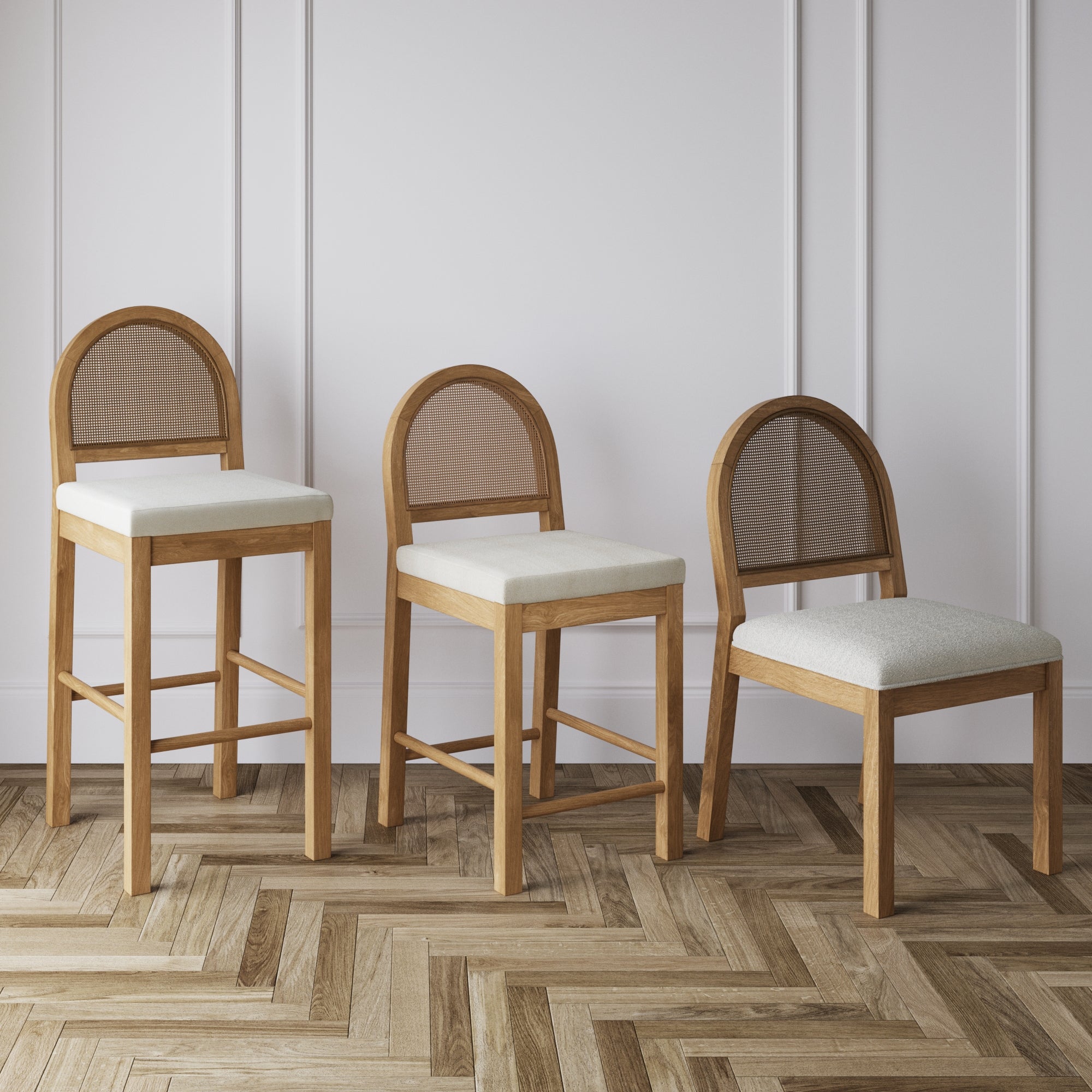 Three wooden chairs with light beige cushions are arranged side by side, each at a different height, against a white paneled wall on a herringbone-patterned wood floor.