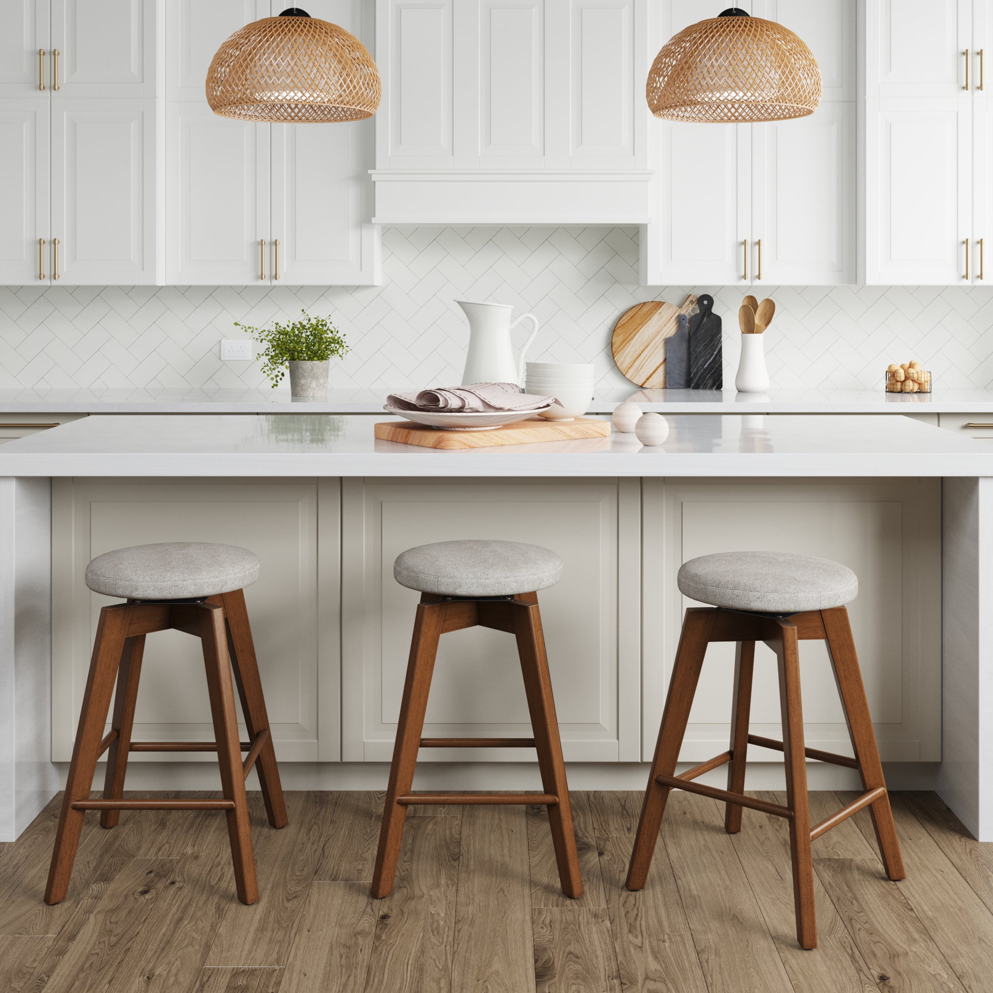 A modern kitchen with white cabinets, a white island, three wooden stools with gray cushions, wooden cutting boards, a pitcher, and two woven pendant lights. The floor is wood and there are small plants on the counter.