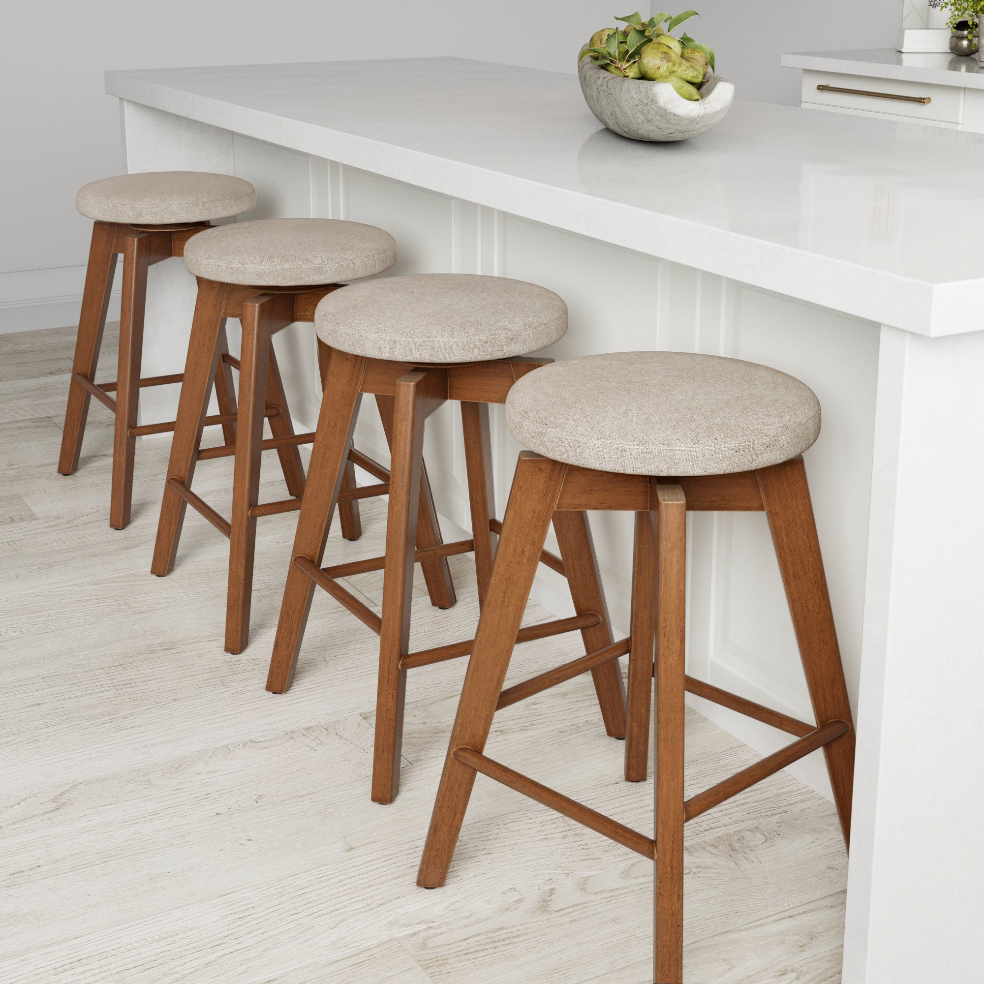 Four wooden bar stools with round, beige upholstered seats are lined up against a white kitchen island. A decorative bowl with green apples sits on the countertop. The floor is light wood.