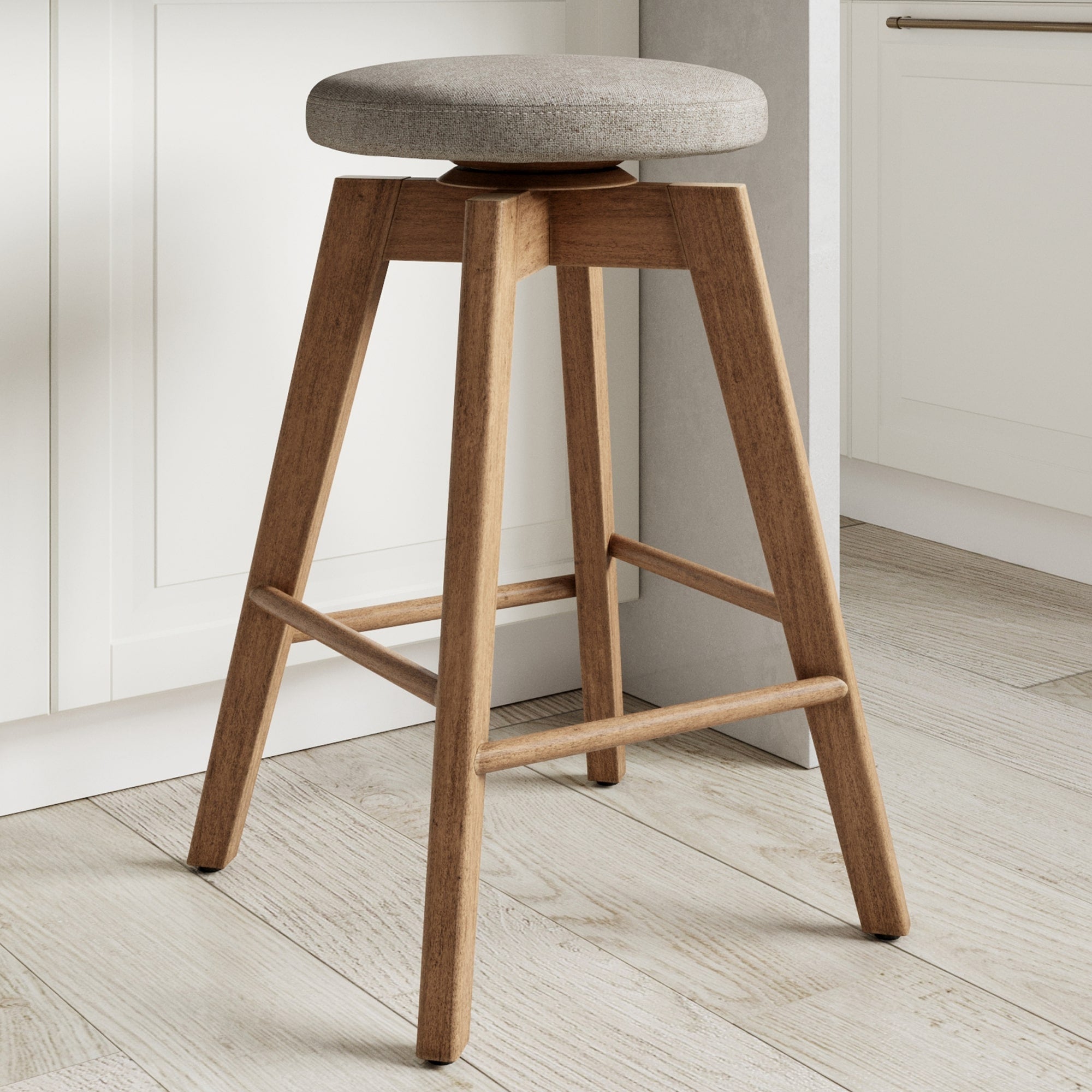 A wooden barstool with four angled legs, cross supports, and a round cushioned seat upholstered in light grey fabric, placed on a light-colored wood floor in a modern kitchen setting.