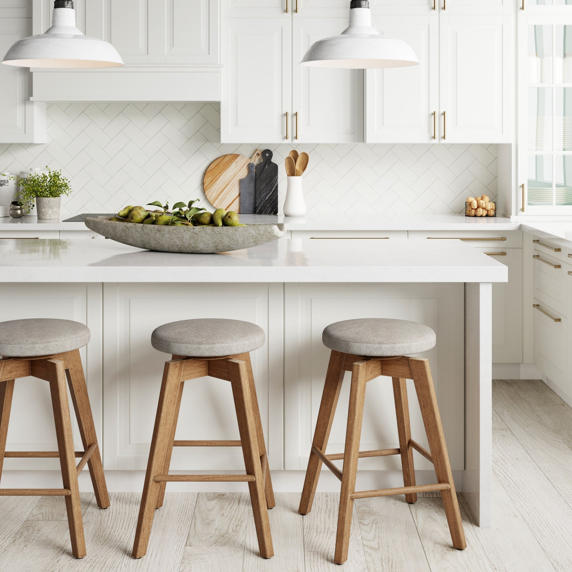 A bright, modern kitchen with white cabinets, a white island, and three Nathan James Wood Swivel Cushion Counter Height Bar Stools in brown with gray seats and 360° swivel. Pendant lights illuminate a bowl of green pears on the counter.