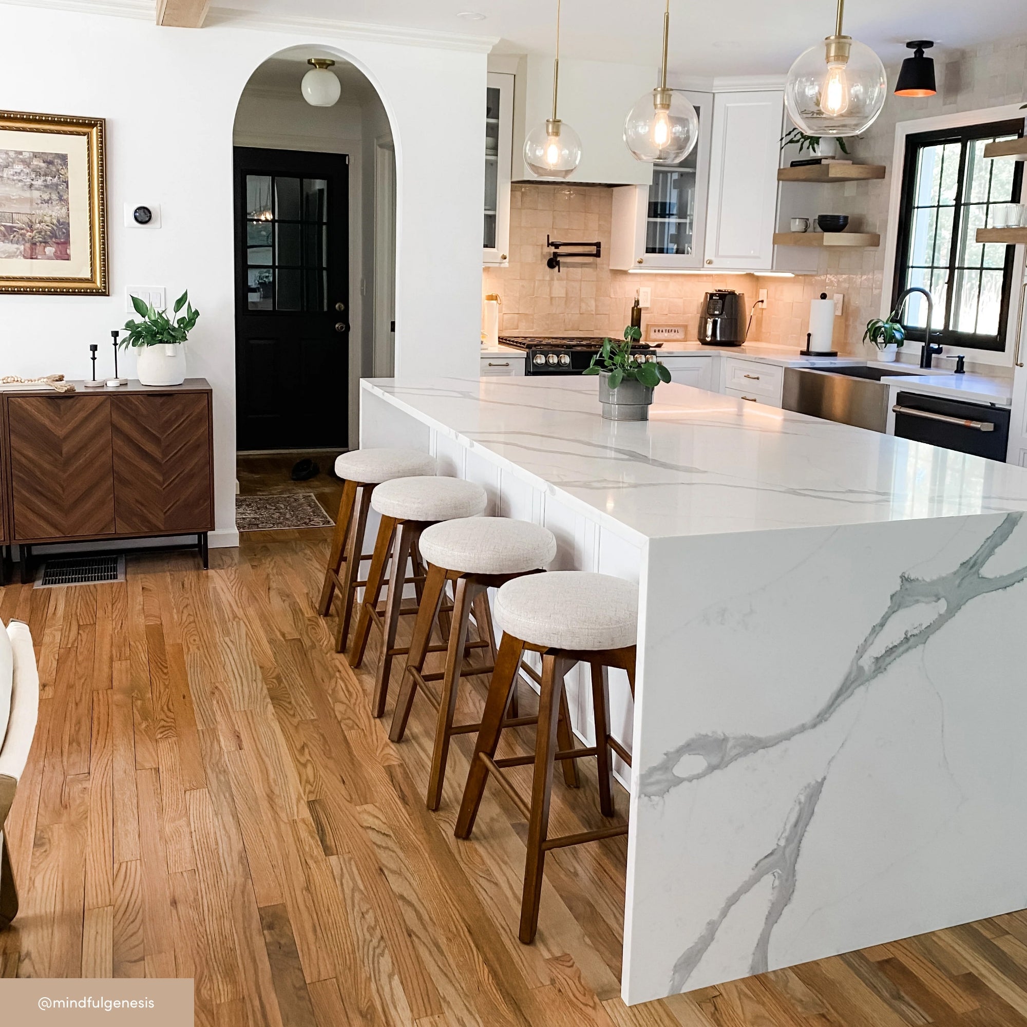 Modern kitchen with a large white marble island, four cushioned bar stools, wooden floors, white cabinets, pendant lights, plants, and a black door in the background.