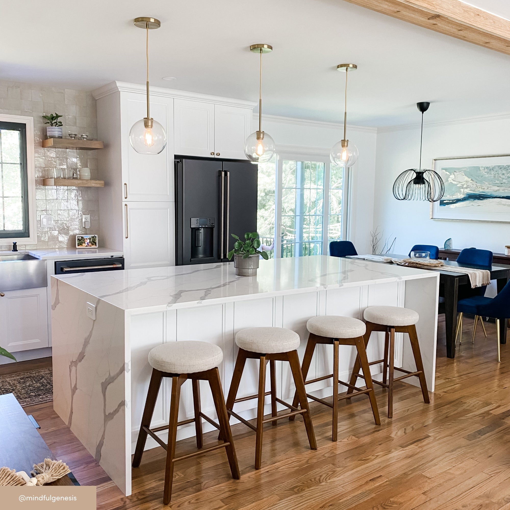 A modern kitchen with a white marble island, four cushioned wooden stools, pendant lights, a black fridge, white cabinets, and a dining area with a large painting and blue chairs in the background.