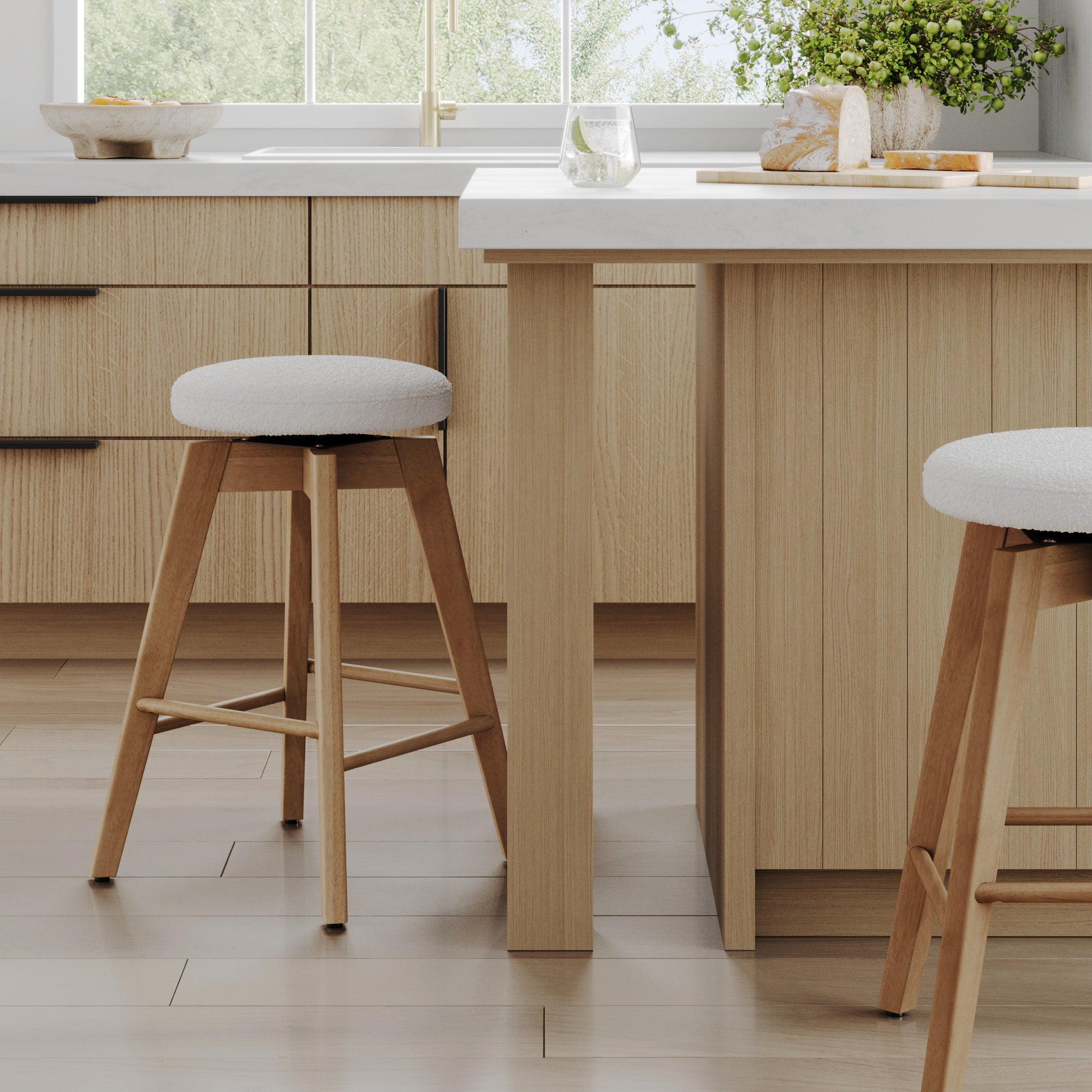 A modern kitchen with light wood cabinets and an island, two wooden stools with white cushions, a glass of water, a cutting board with bread, and a bowl of fruit, with sunlight streaming through a window.