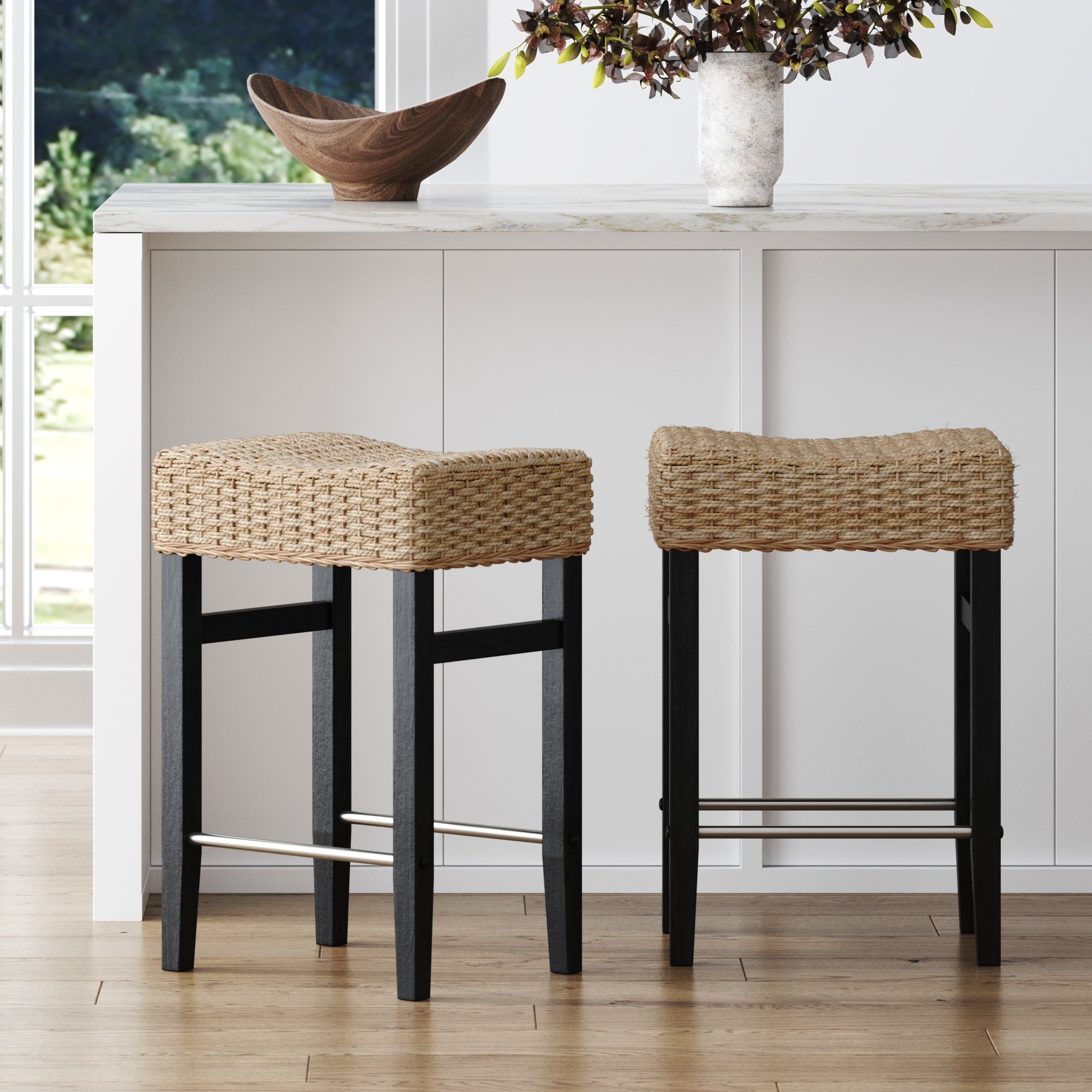 Two Nathan James Seagrass Counter Height Bar Stools in black sit at a white kitchen island, decorated with a wooden bowl and a vase of branches. Large windows fill the space with natural light.