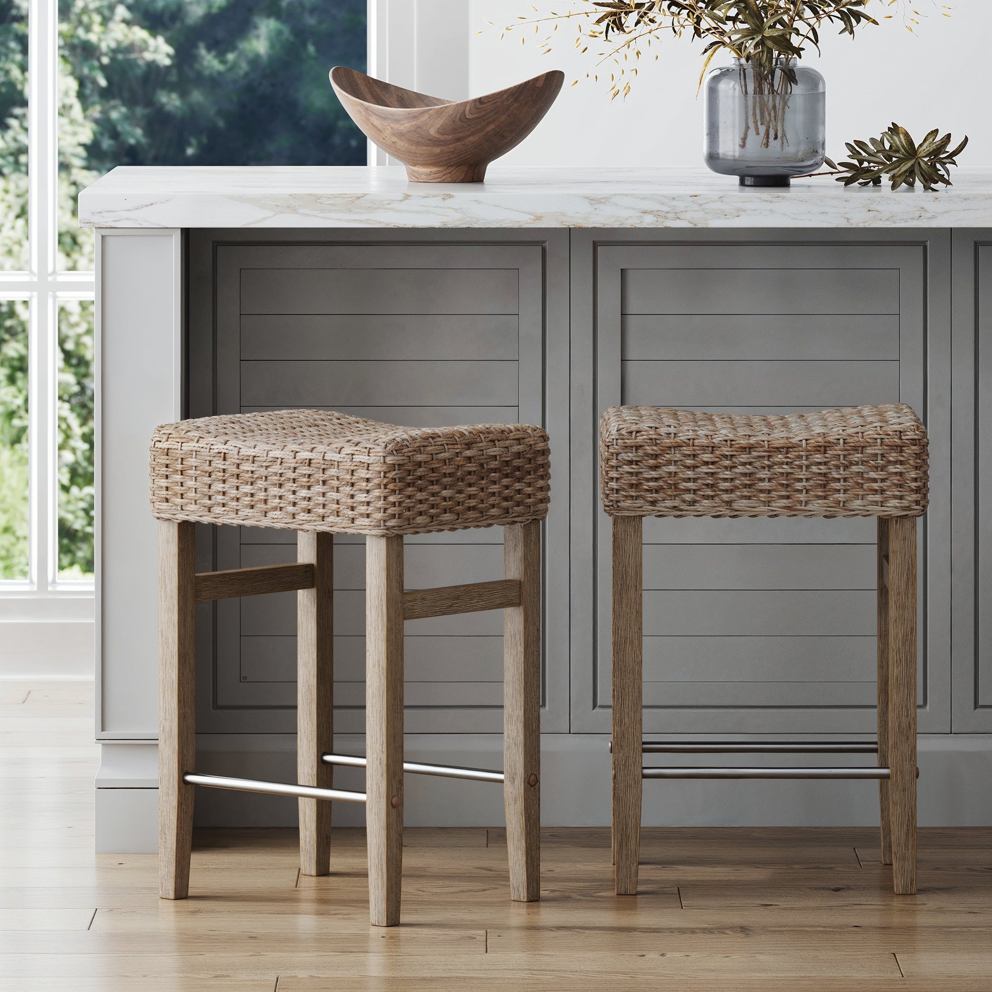 Two Nathan James Seagrass Counter Height Bar Stools in brown stand by a gray kitchen island with a white marble countertop, accented by a wooden bowl and vase of foliage. Large windows overlook lush greenery outside.