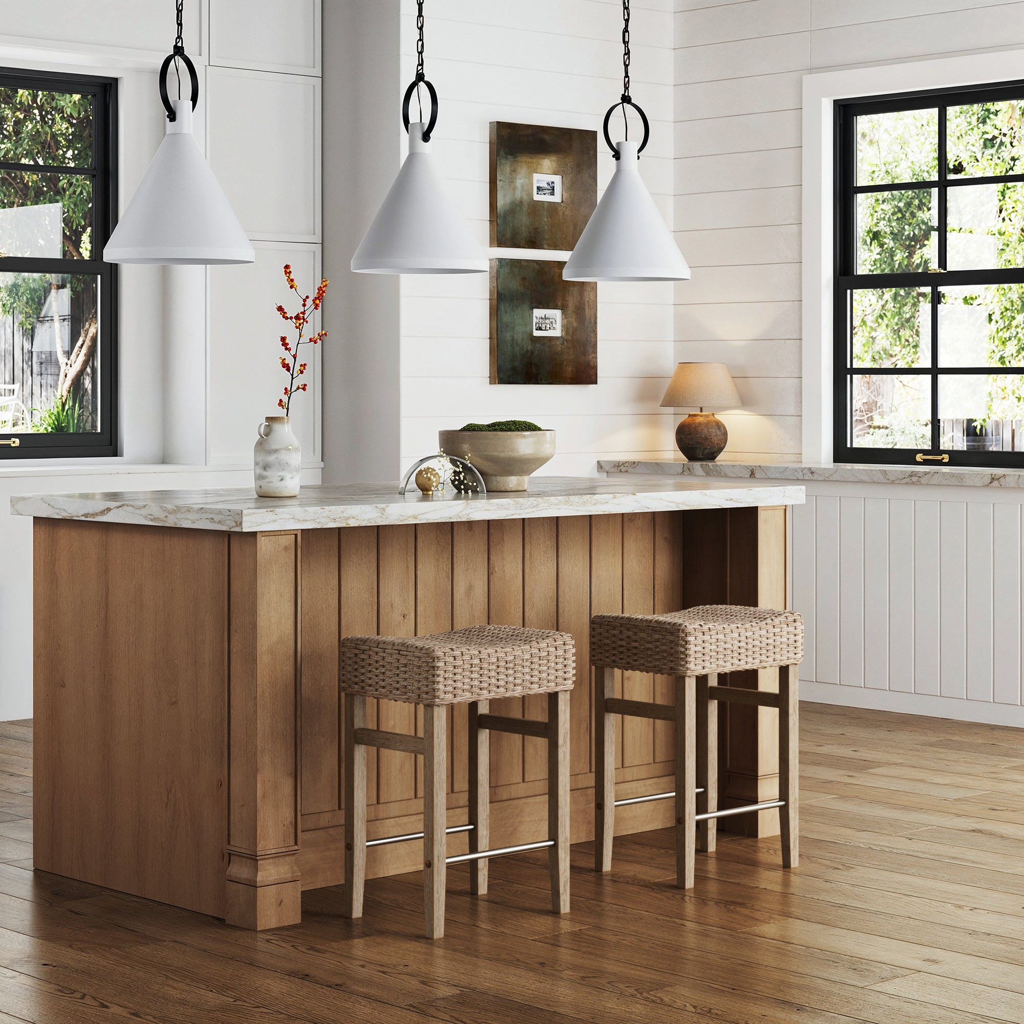 A modern kitchen with a wooden island, two Nathan James Seagrass Counter Height Bar Stools in brown, a marble countertop, pendant lights, decorative vases, framed wall art, and large windows letting in natural light.