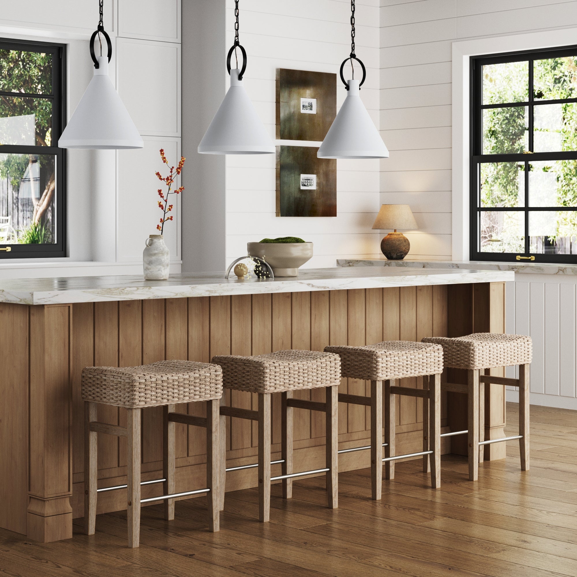 A modern kitchen with a wooden island, four Nathan James Woven Seagrass Counter Height Bar Stools in brown, white pendant lights, marble countertop, decorative vases, framed wall art, and large windows letting in natural light.
