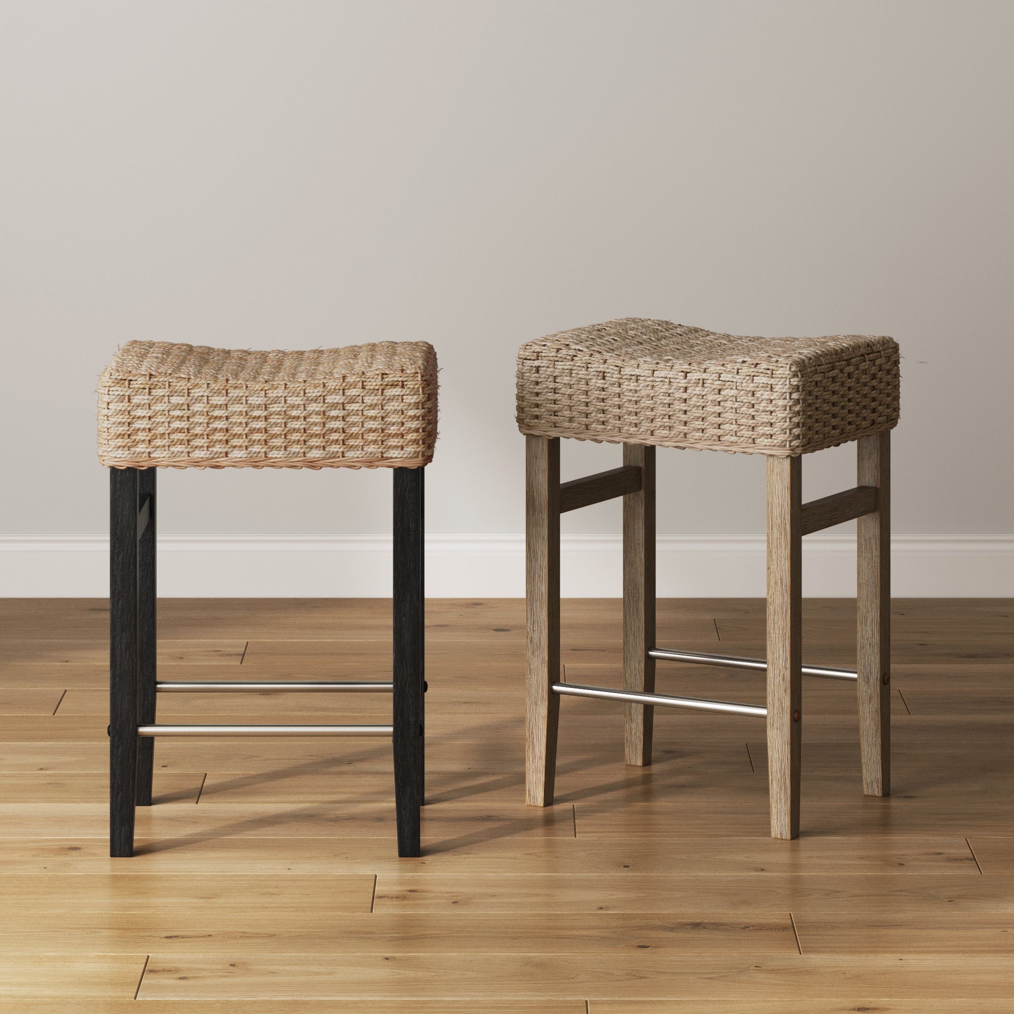 Two woven stools with saddle-shaped seats sit side by side on a wooden floor. One has black legs, the other has light wood legs. A plain white wall is in the background.