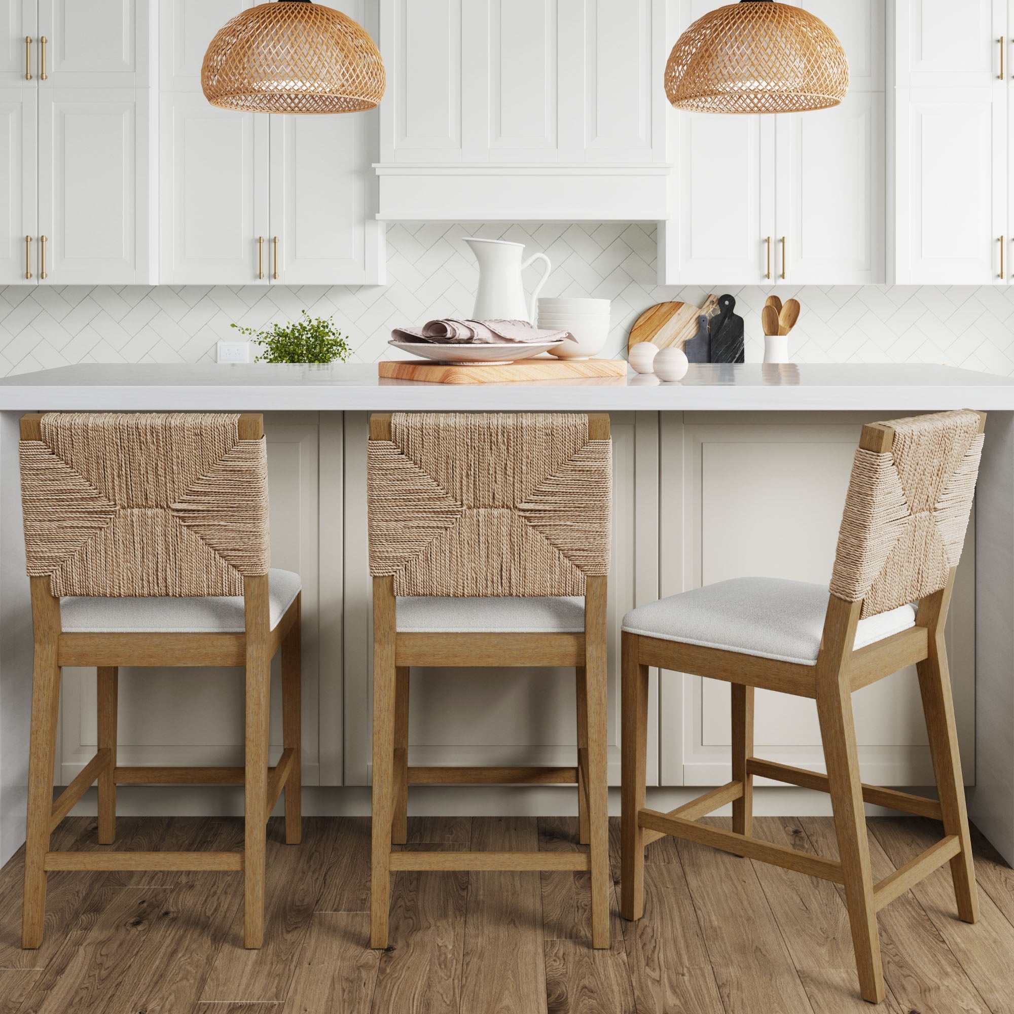Three Nathan James Seagrass Counter Height Bar Stools in light brown are lined up at a white kitchen island with white cabinets, wood accents, and two wicker pendant lights above.