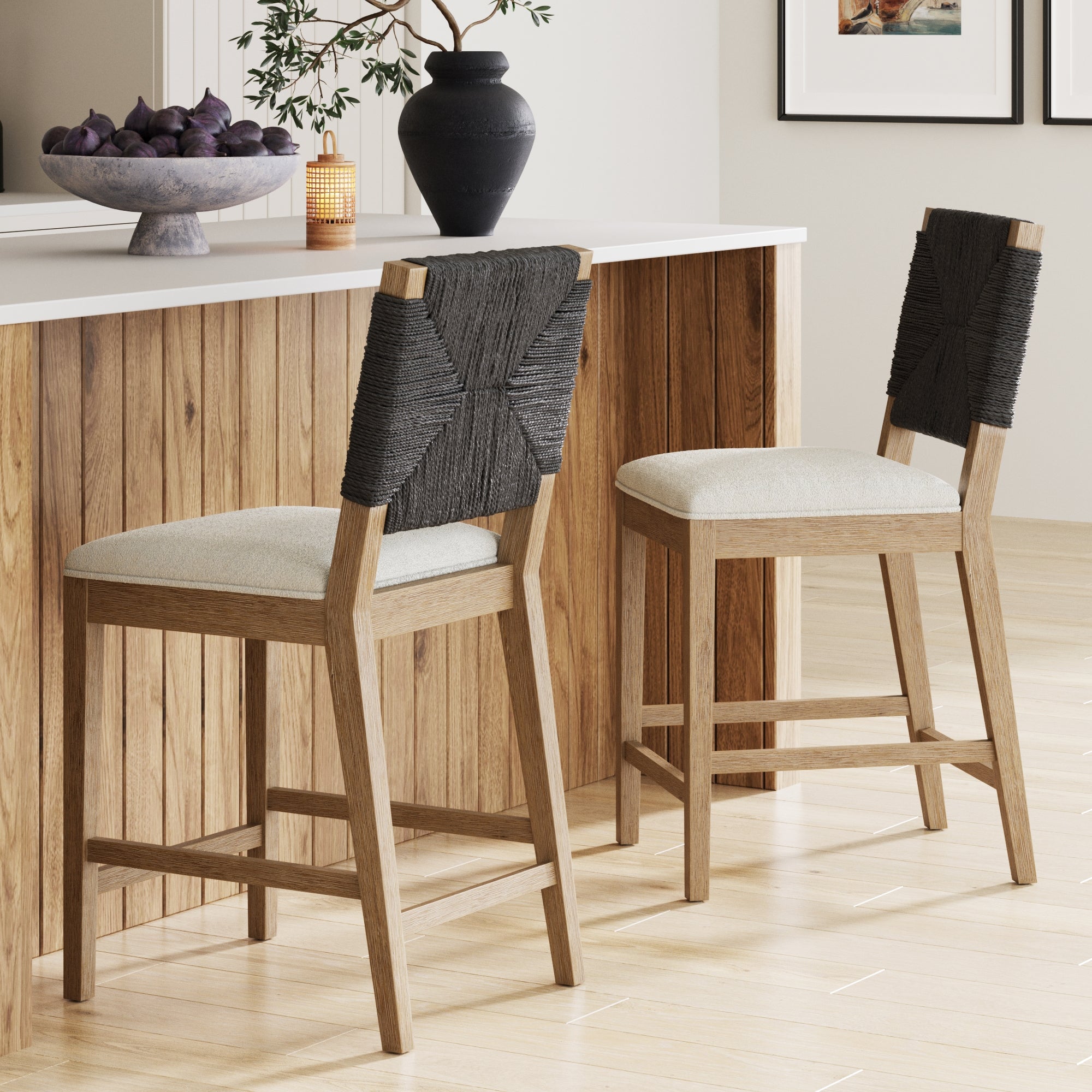 Two Nathan James Boucle & Seagrass Counter Height Bar Stools (Set of 2) with black woven backs and beige cushions are at a wooden kitchen island. A bowl of fruit, vase, and framed pictures add a modern, cozy touch to the space.