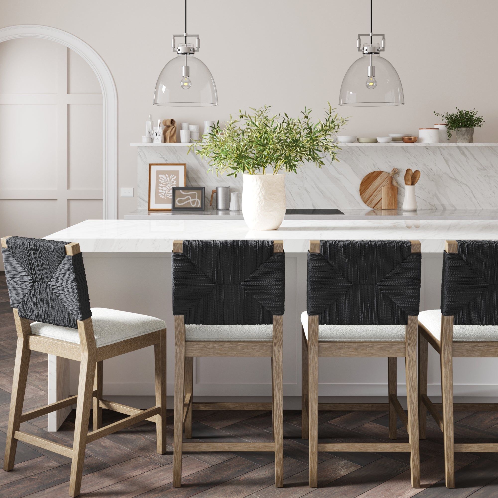 Modern kitchen featuring a marble island, a set of 4 Nathan James Boucle & Seagrass Counter Height Bar Stools in black with cream cushions, pendant lights, a vase with green branches, and minimalist decor on the counter and wall.