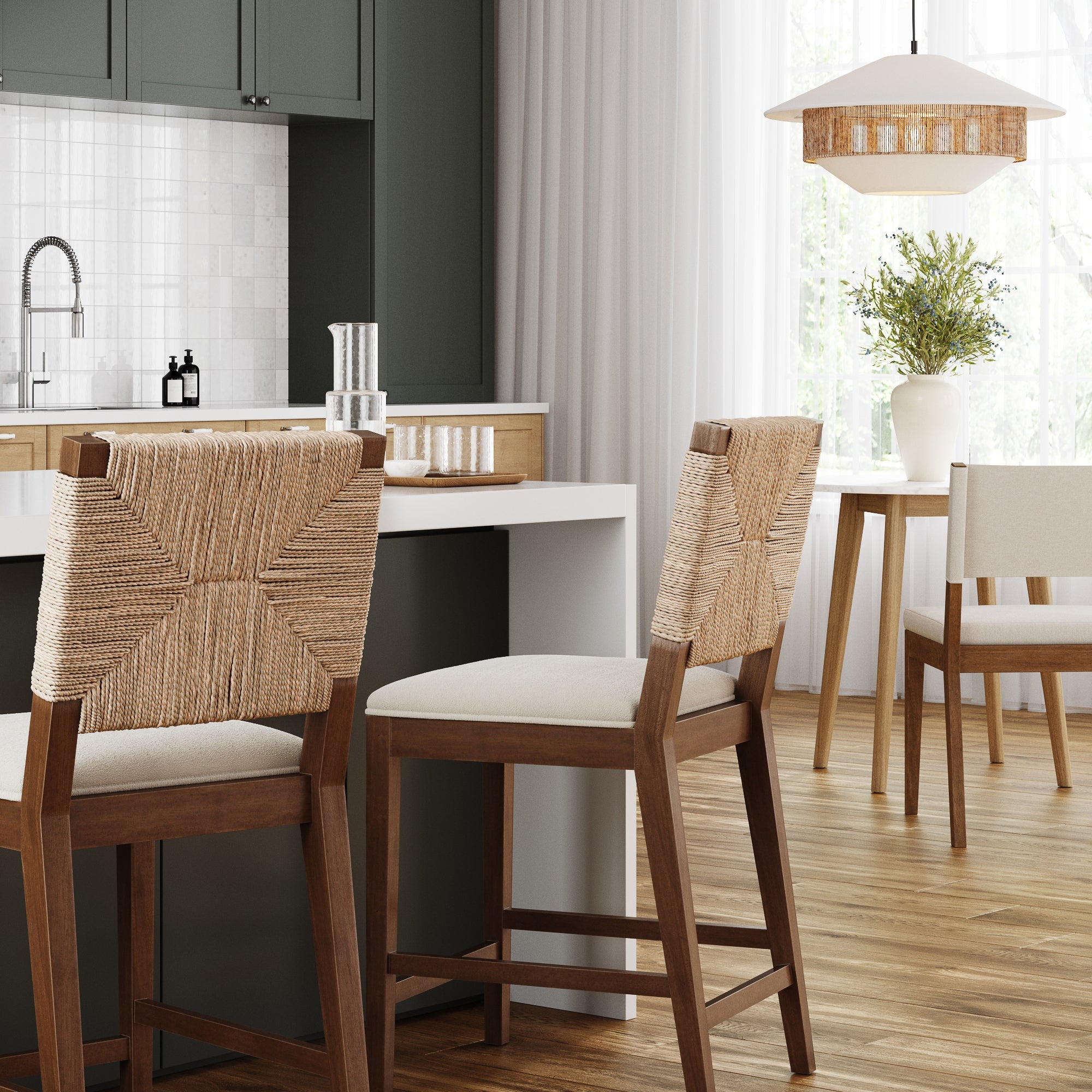 Modern kitchen with a white island, two woven-back barstools, wood flooring, a small dining table with a vase of greenery, and soft natural light filtering through sheer curtains.