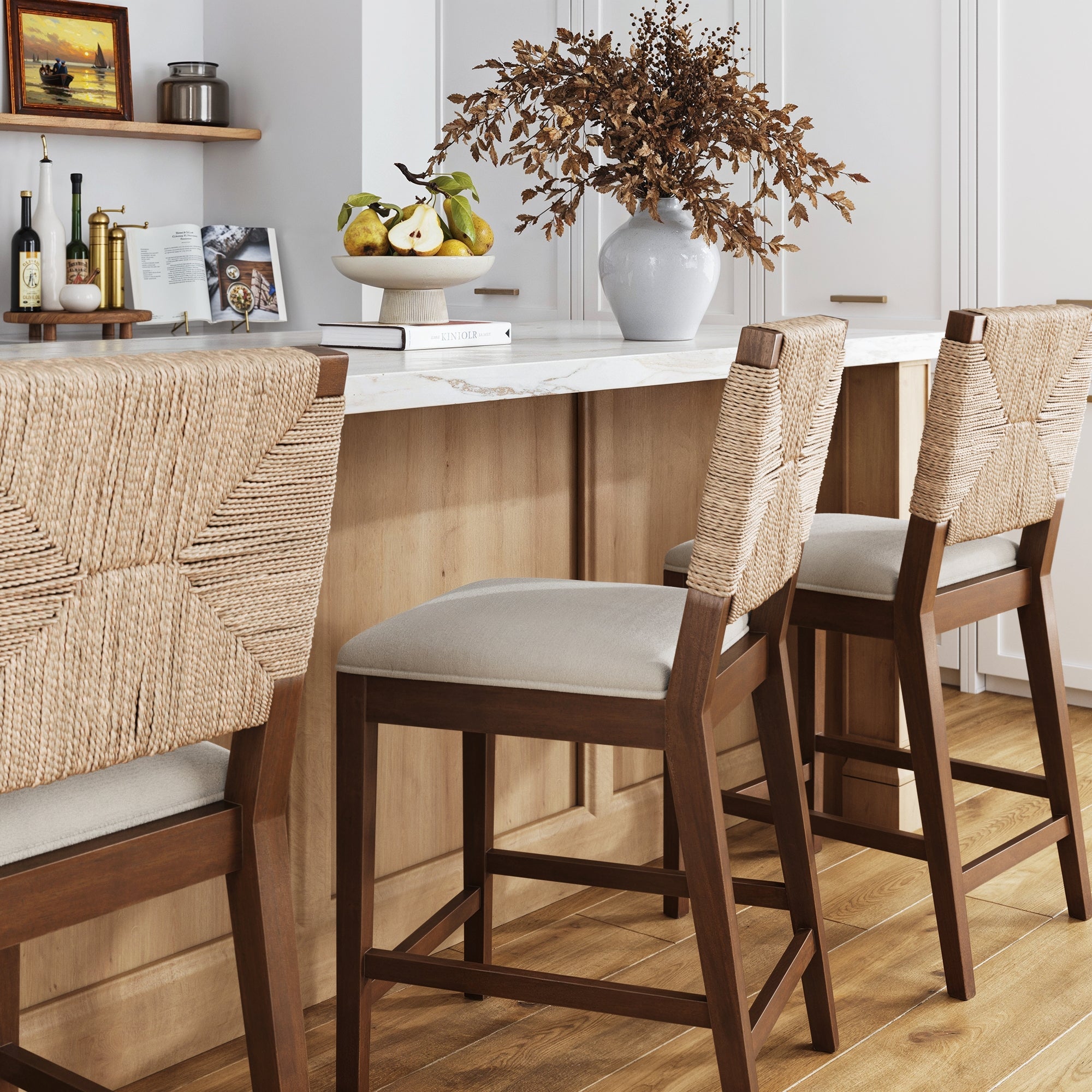 Three Nathan James Seagrass Counter Height Bar Stools in dark brown (set of 3) with woven backs and cushioned seats are arranged at a marble kitchen island, accented by light wood floors, cabinetry, a vase with dried branches, and a fruit bowl.