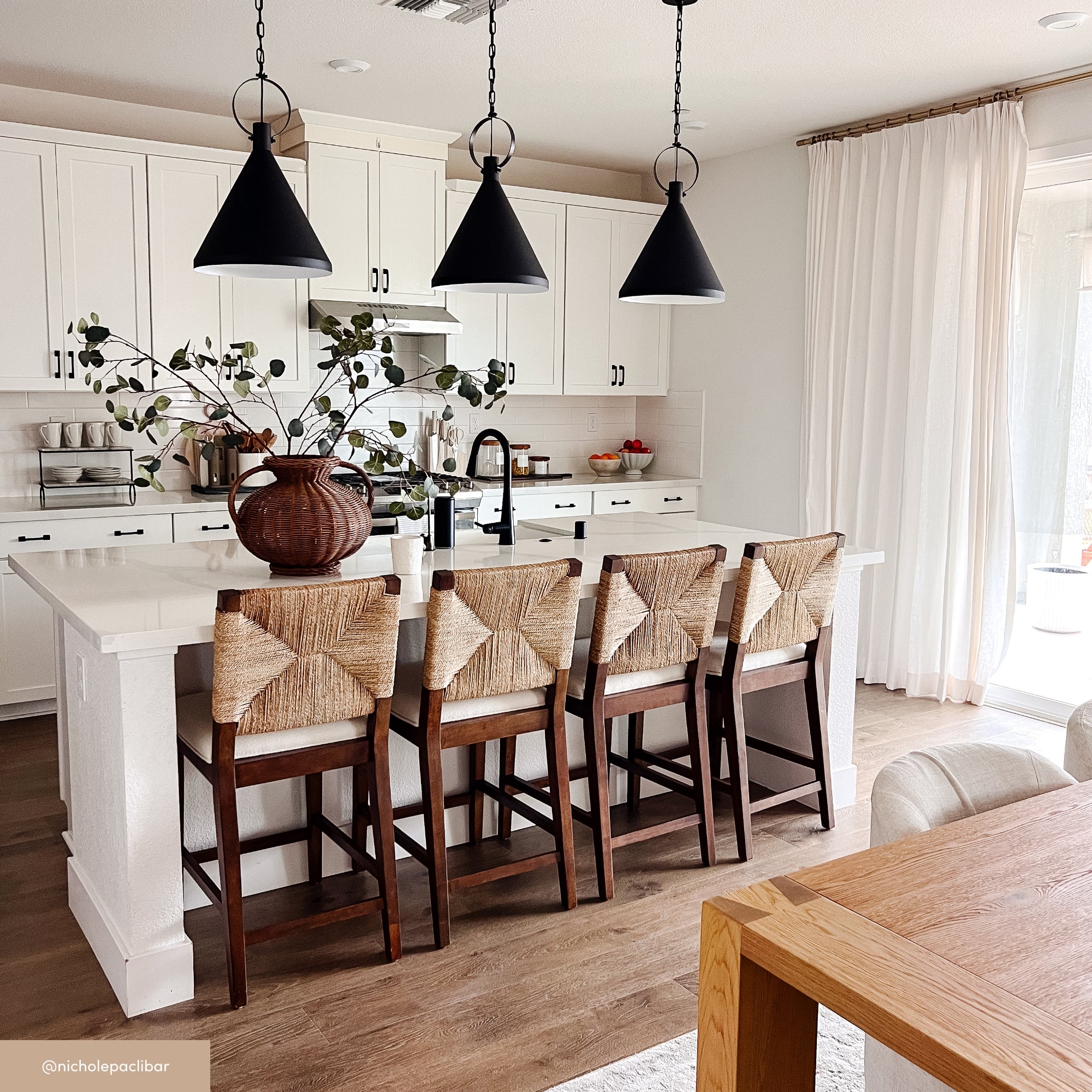 Bright, modern kitchen with white cabinets, black pendant lights, and a large island with four wooden barstools featuring woven backs. A brown vase with greenery sits on the island near a black faucet.