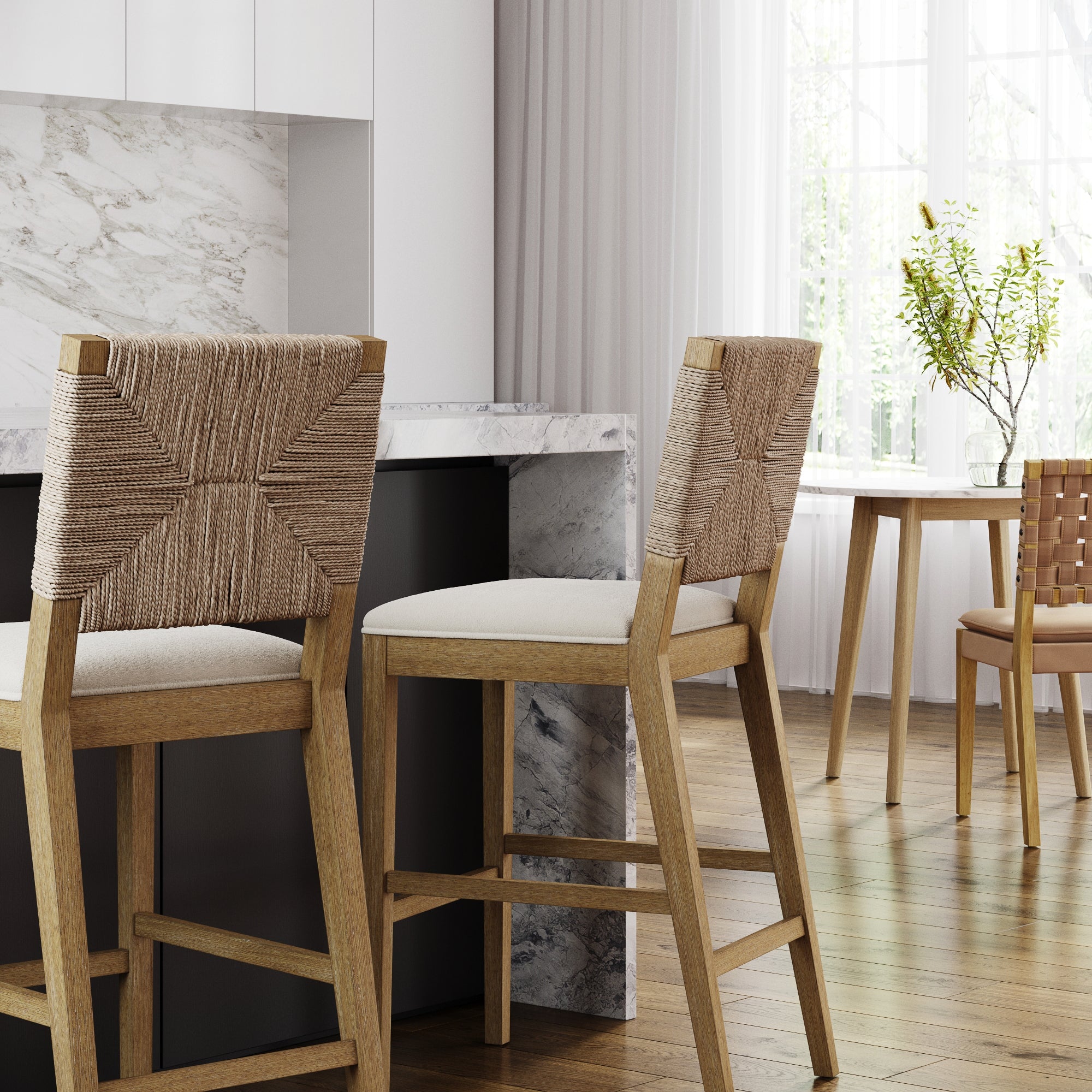 Two Nathan James Boucle & Seagrass Bar Stools in light brown are placed at a marble kitchen island. Sunlight warms a wooden dining table with a vase of branches in the background.