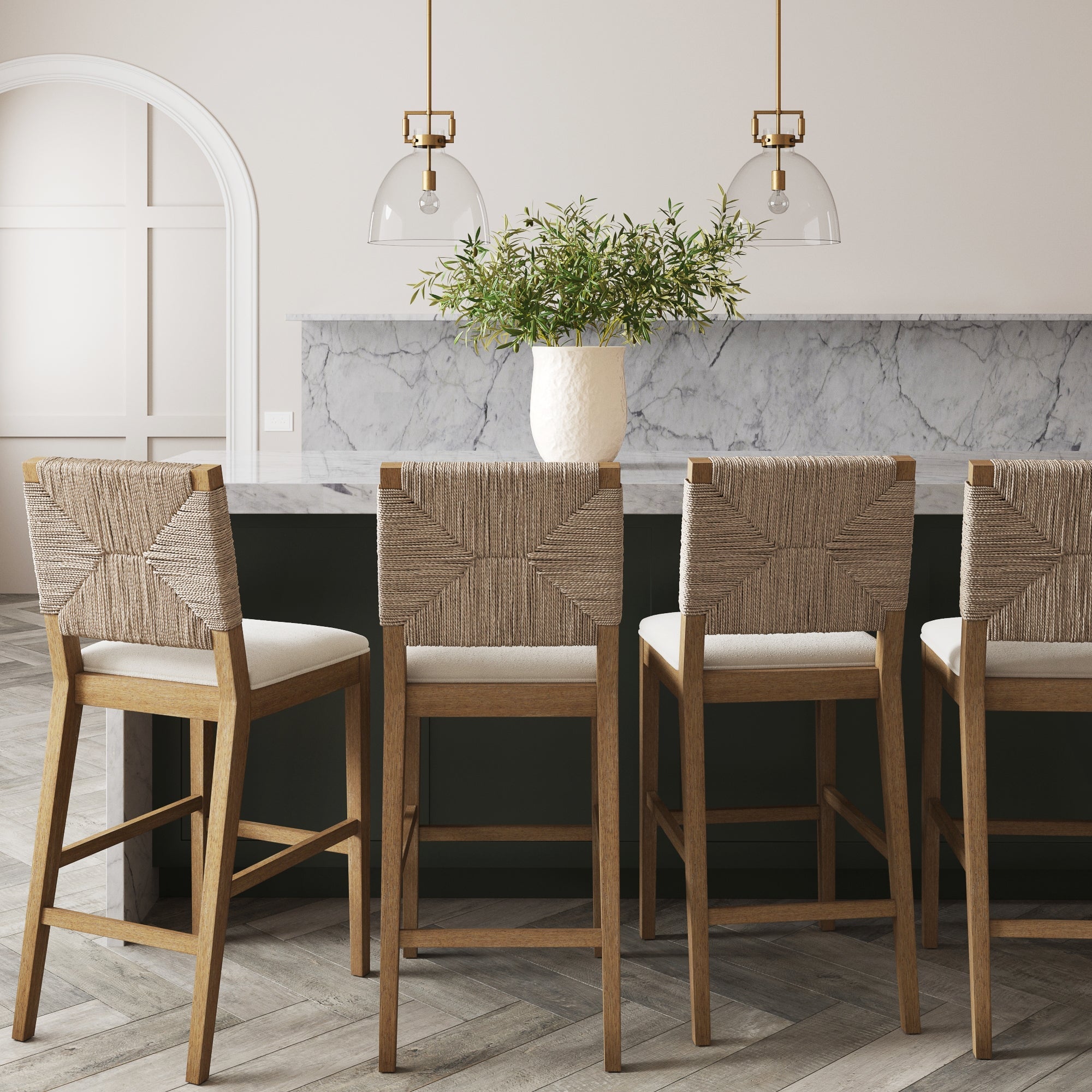 Four Nathan James Boucle & Seagrass Bar Stools in light brown are lined up at a marble island. Two glass pendant lights hang above, and a white vase with green foliage sits on the counter in this modern, neutral-toned space.