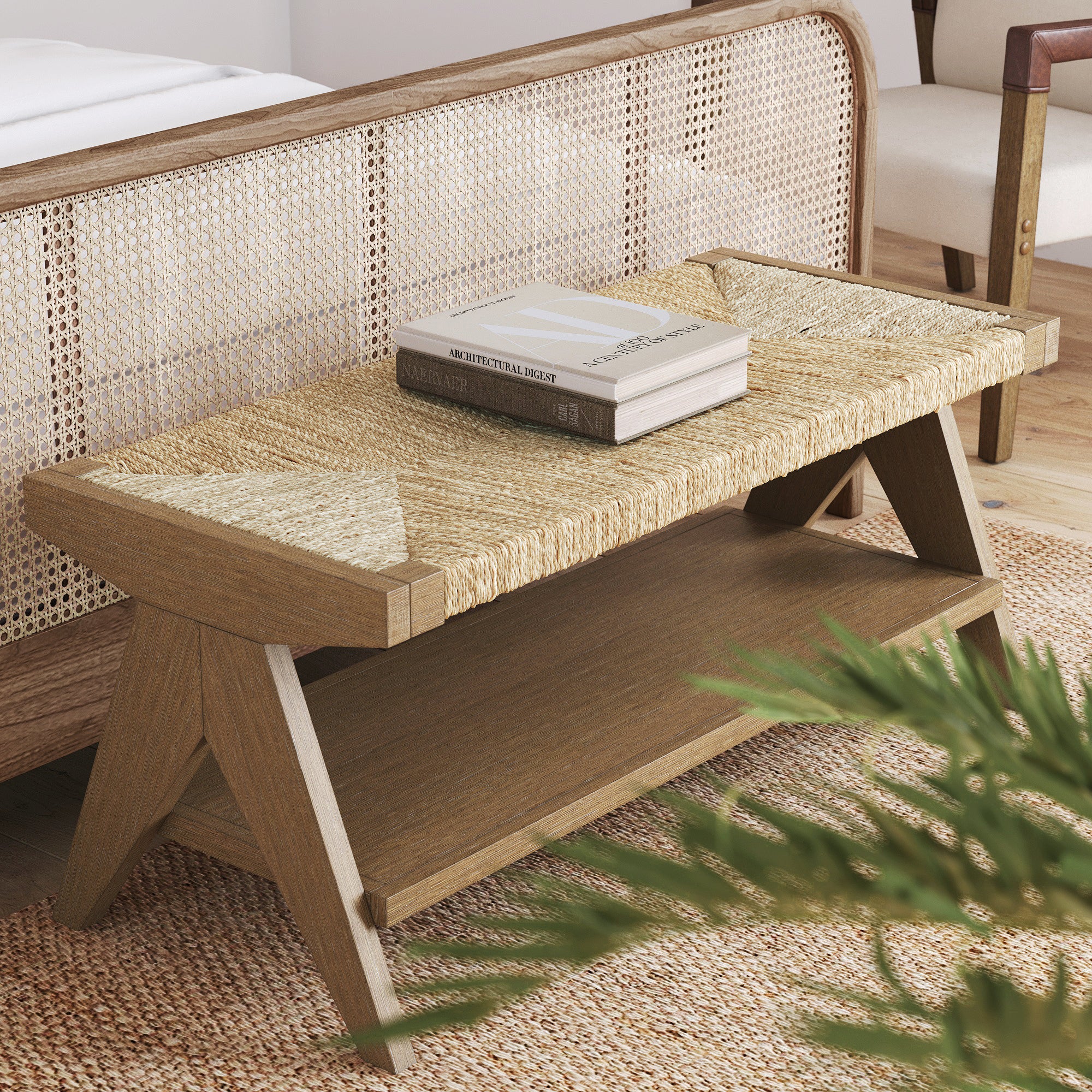 A wooden bench with a woven seat and lower shelf stands at the foot of a rattan bed. Two books rest on top of the bench. A plant is partially visible in the foreground on a woven rug.