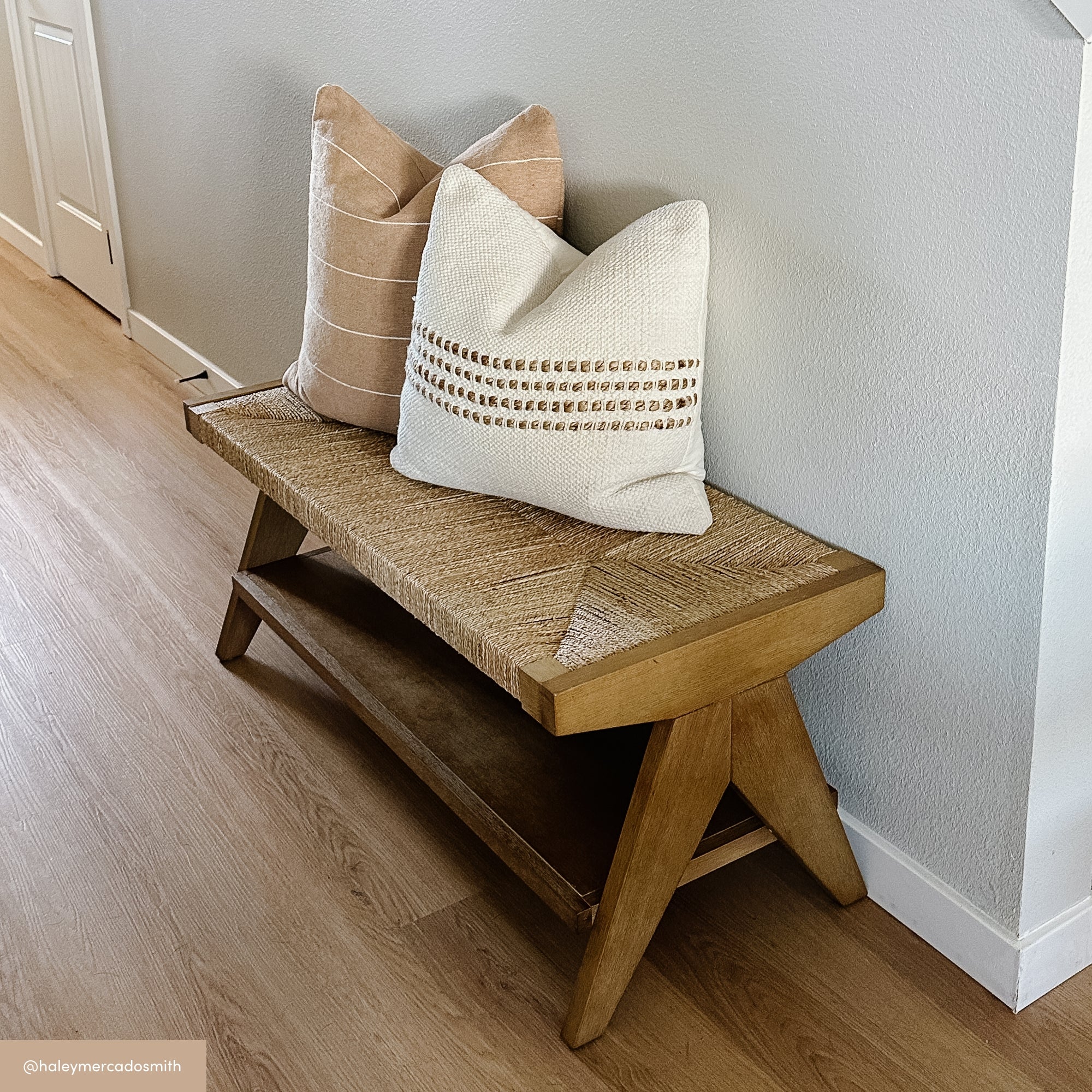 A wooden bench with a woven seat sits against a light gray wall. Two neutral-colored throw pillows, one striped and one with woven detailing, rest on the bench. The floor is light wood.