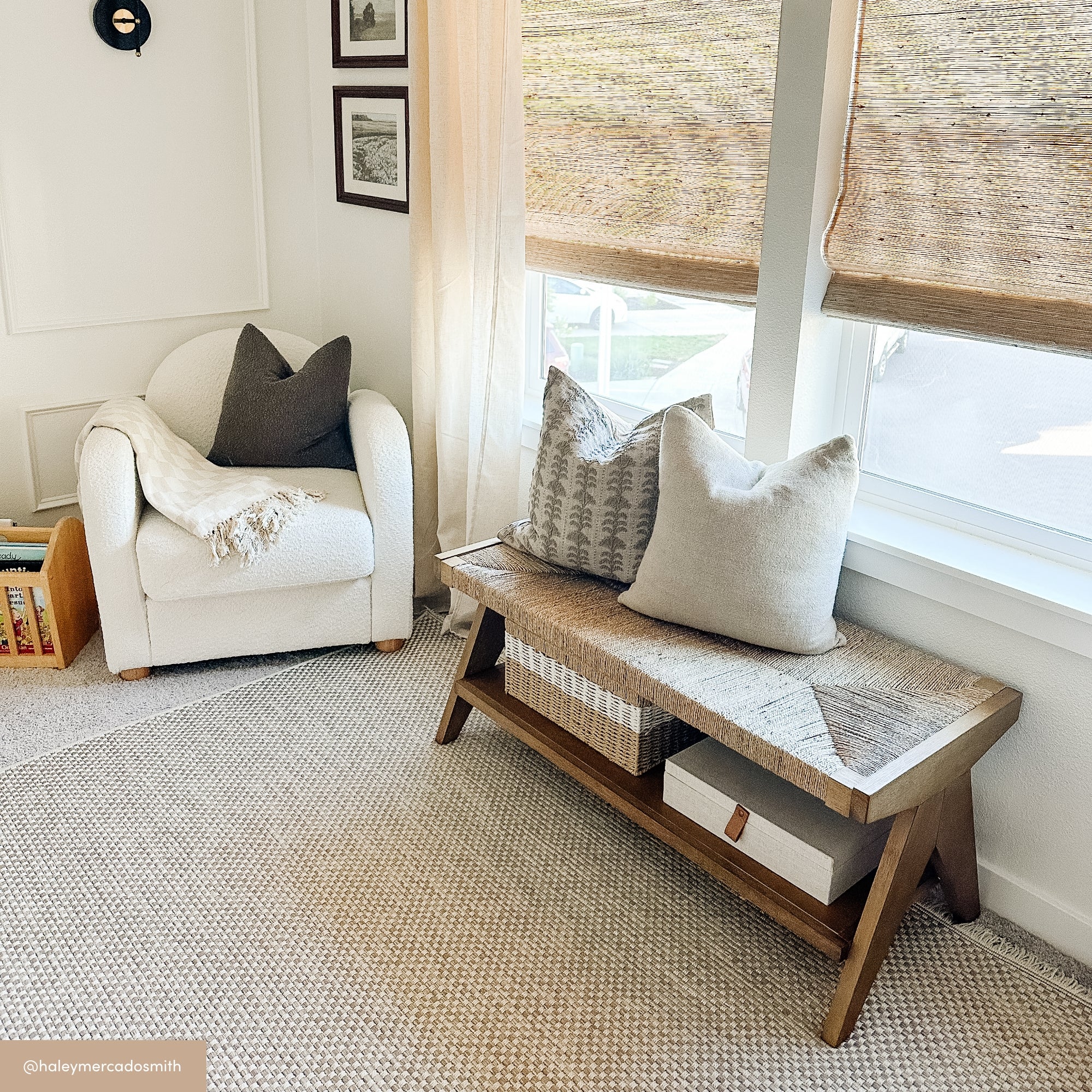 A cozy corner with a white armchair, a beige throw blanket, framed pictures on the wall, a wooden bench with decorative pillows, woven blinds, and a carpeted floor. Sunlight filters in through the window.