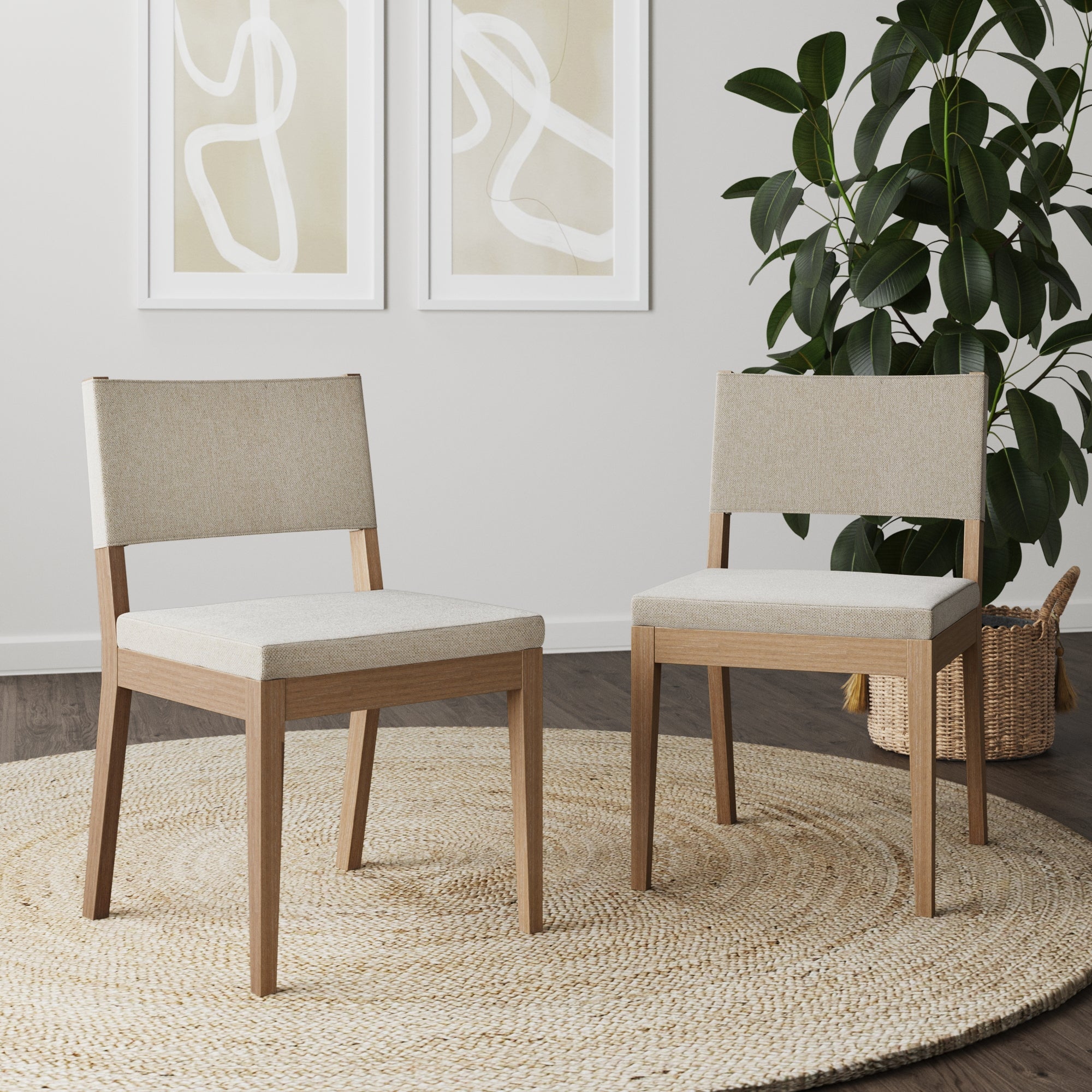 Two Nathan James Upholstered Dining Chairs in light brown sit on a round woven rug. Behind them, a large leafy plant in a basket and two abstract art prints decorate the white wall.