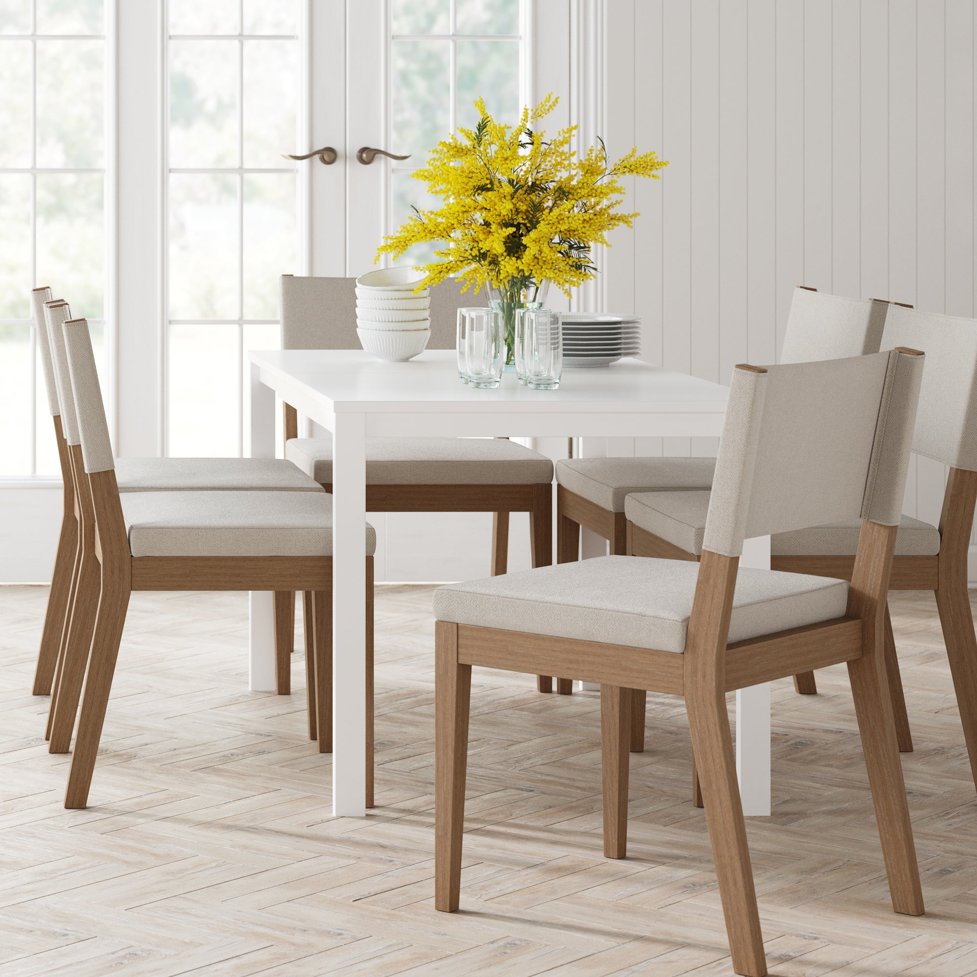 A modern dining room with a white table and six Nathan James Upholstered Dining Chairs in light brown. Stacked white plates, glassware, bowls, and a vase of yellow flowers sit atop the table. Large windows in the background fill the room with natural light.