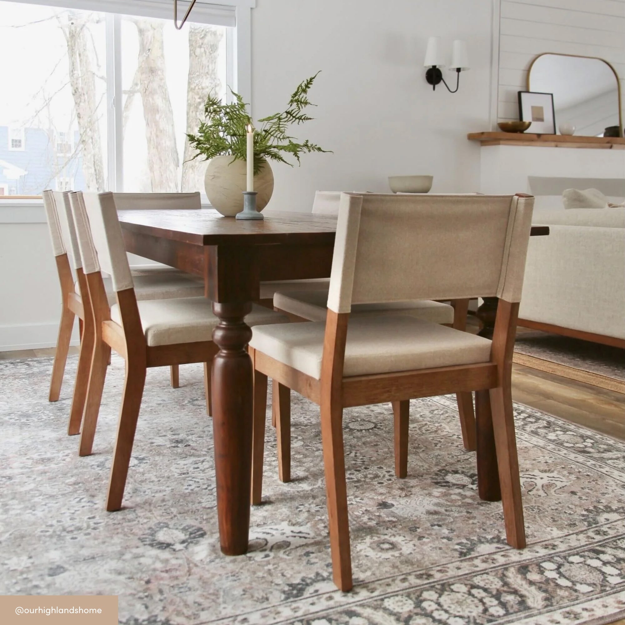 A dining room with a wooden table and four upholstered chairs, a vase with greenery on the table, a patterned rug underneath, and large windows letting in natural light. A mirror and sofa are visible in the background.