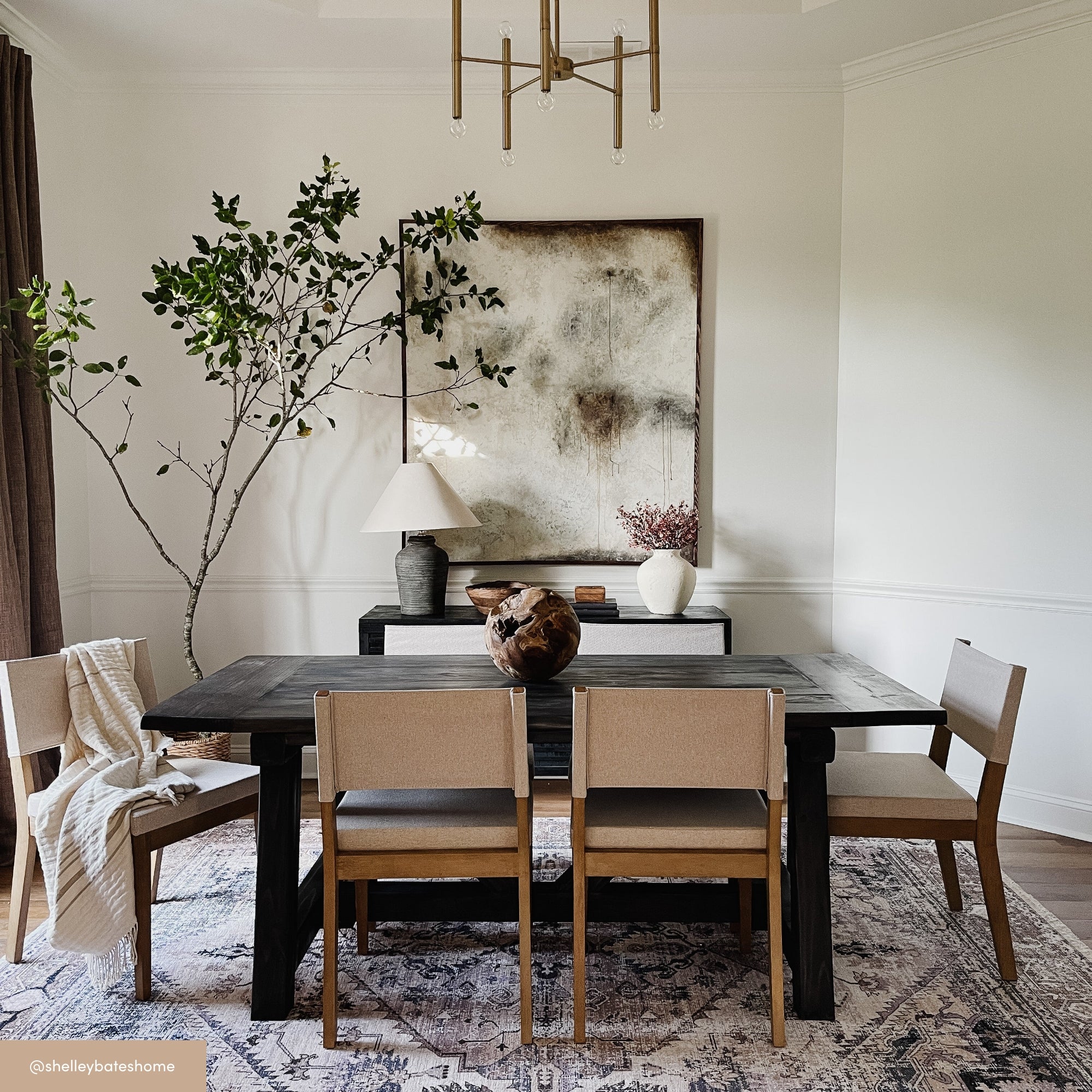 A modern dining room with a black table, beige chairs, a large abstract painting, a sideboard with a lamp, and a tall leafy plant in the corner. A chandelier hangs from the ceiling above a patterned rug.