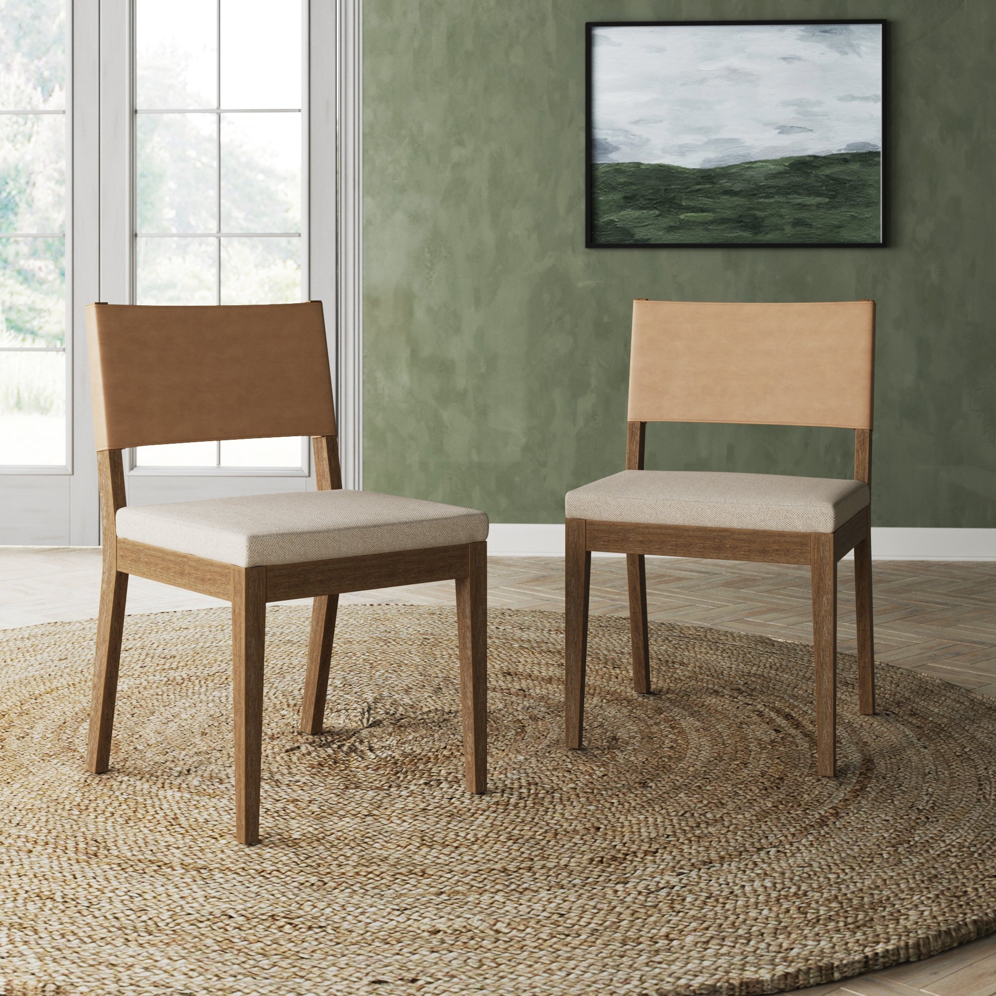 A set of two Nathan James Wood & Faux Leather Dining Chairs in light brown sits on a round woven rug in a green-walled room with a large window and a landscape painting.