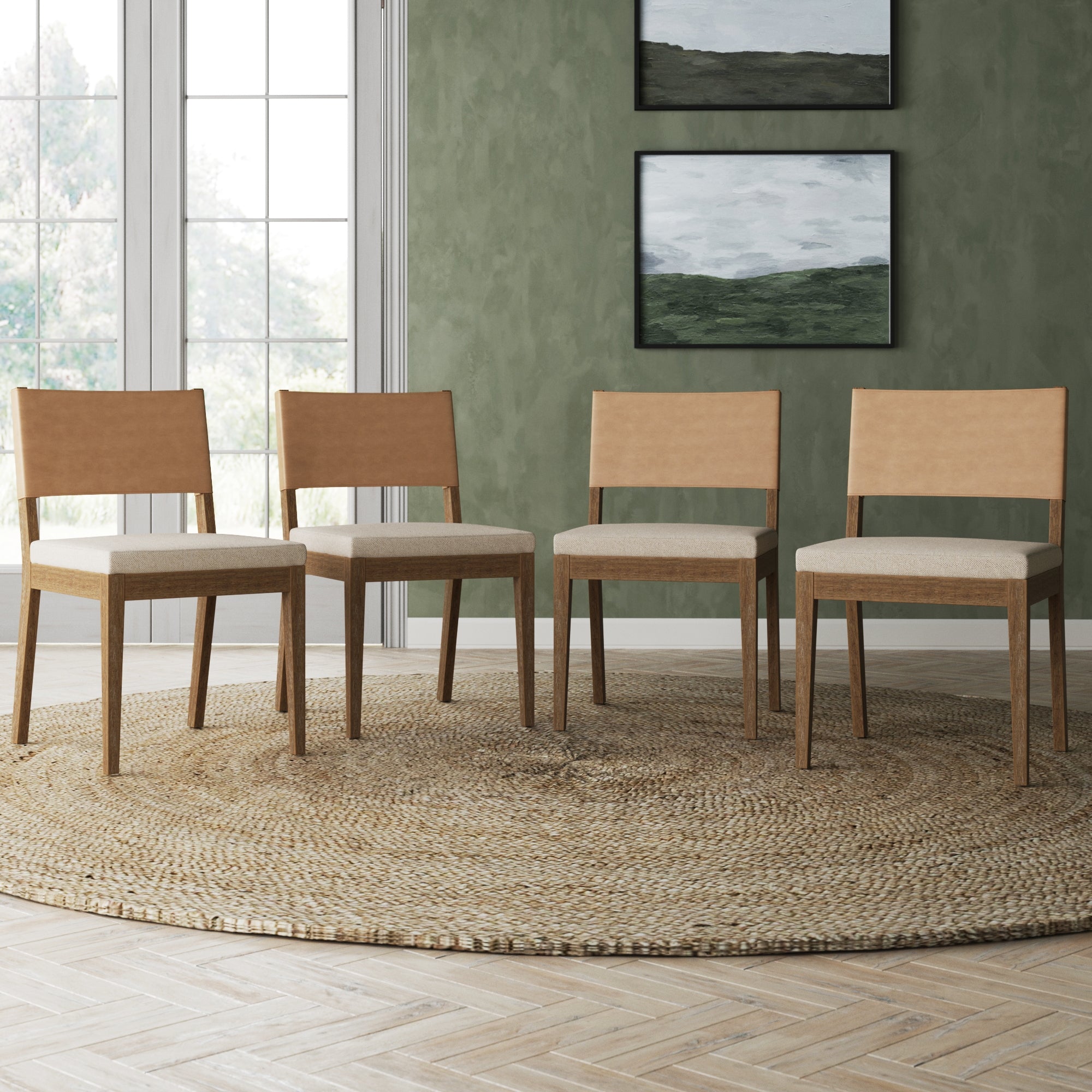 A set of four Nathan James Wood & Faux Leather Dining Chairs in light brown sits on a round woven rug in a room with green walls, two landscape paintings, and large windows that fill the space with natural light.