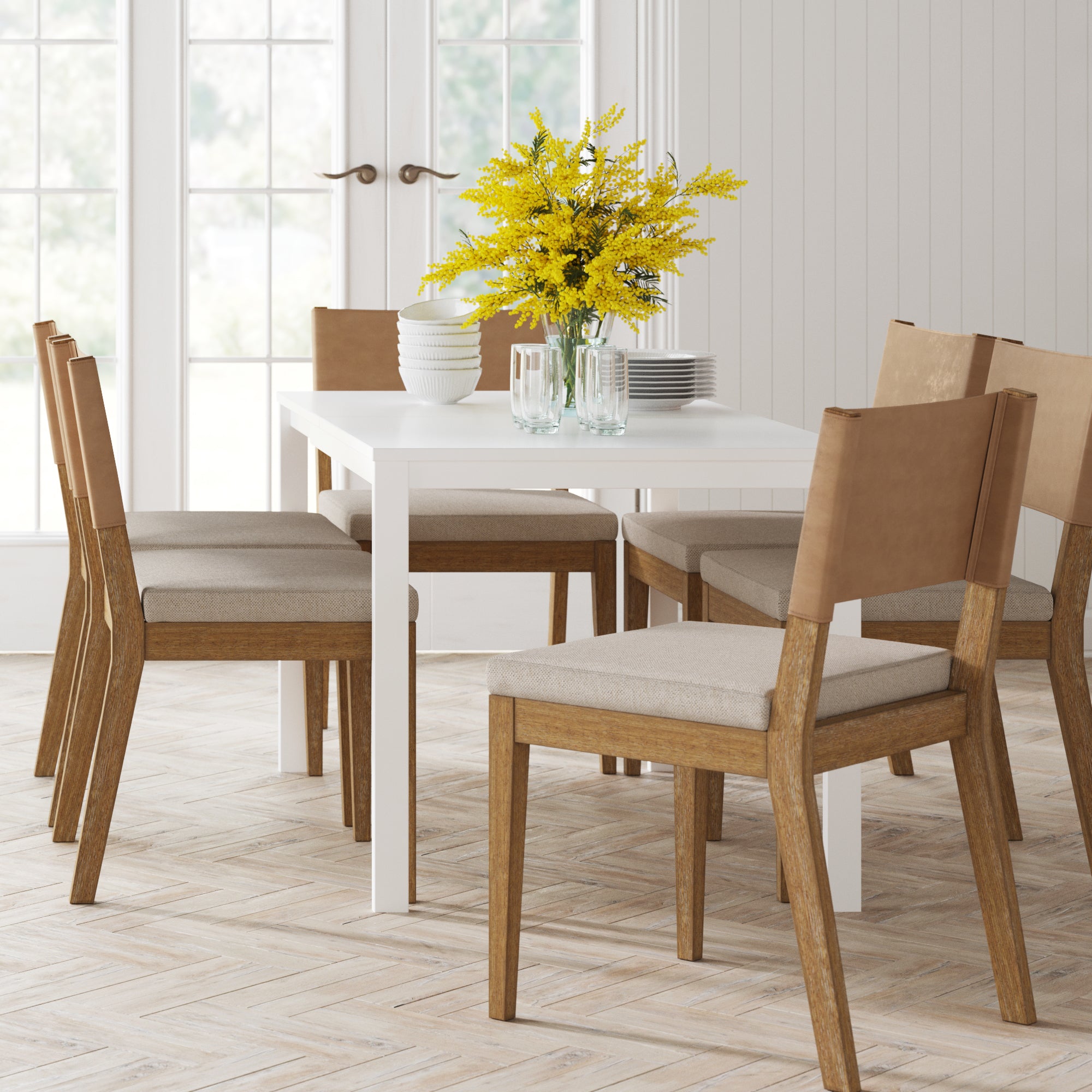A modern dining room features a white table, six Nathan James Wood & Faux Leather Dining Chairs in light brown, and a vase of yellow flowers. Plates, bowls, and glasses are neatly arranged. Large windows and double doors fill the space with light.