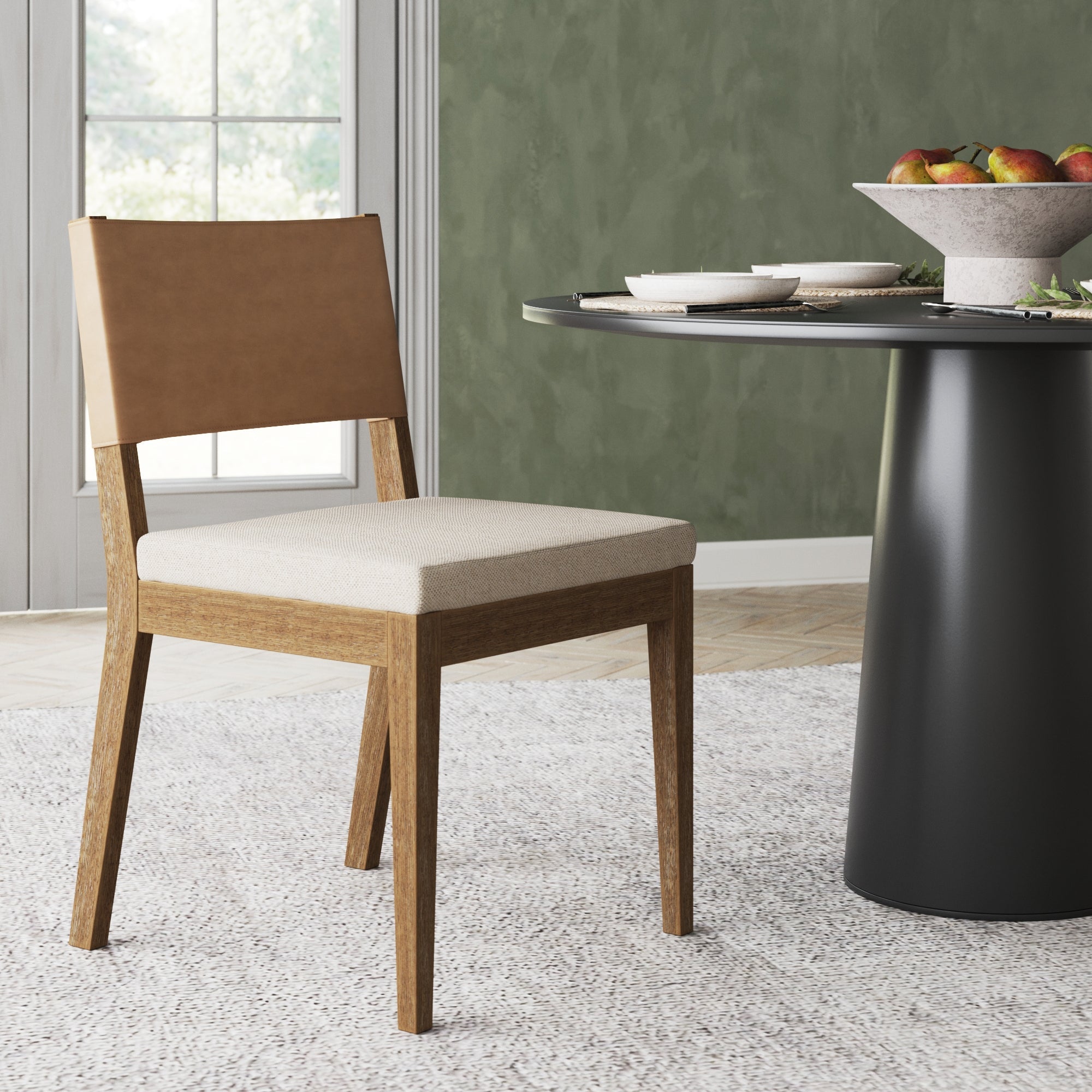 The Nathan James Wood & Faux Leather Dining Chair in light brown, featuring a beige cushioned seat and tan backrest, is shown on a light carpet next to a round black dining table with dishes and fruit, set in a green-walled room with a large window.