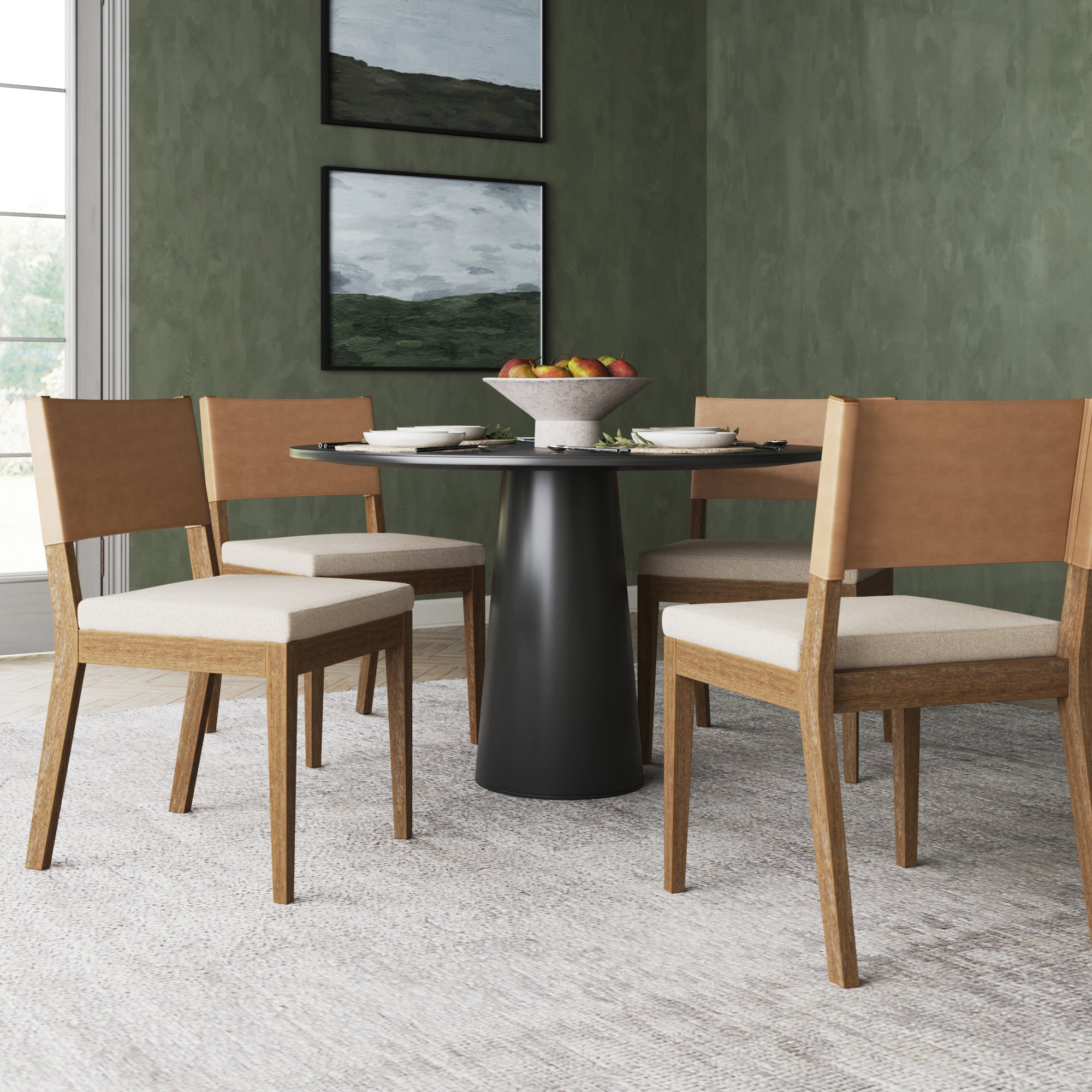 A round black dining table with four wooden chairs sits on a light rug. The table is set with plates and a bowl of fruit. The room has green textured walls and two landscape paintings.