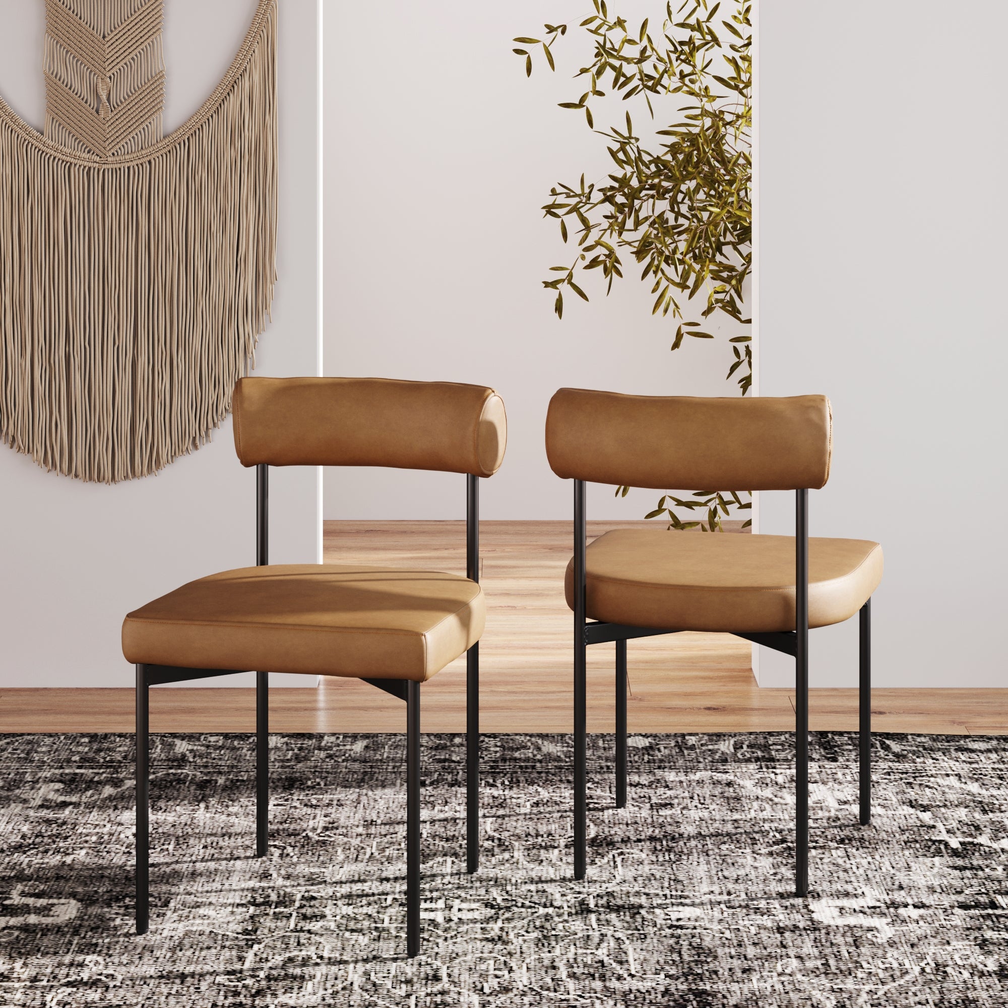 Two Nathan James Faux Leather Modern Dining Chairs (Set of 2) in tan with black metal legs are placed on a dark patterned rug in a bright room, with a woven wall hanging and leafy plant in the background.
