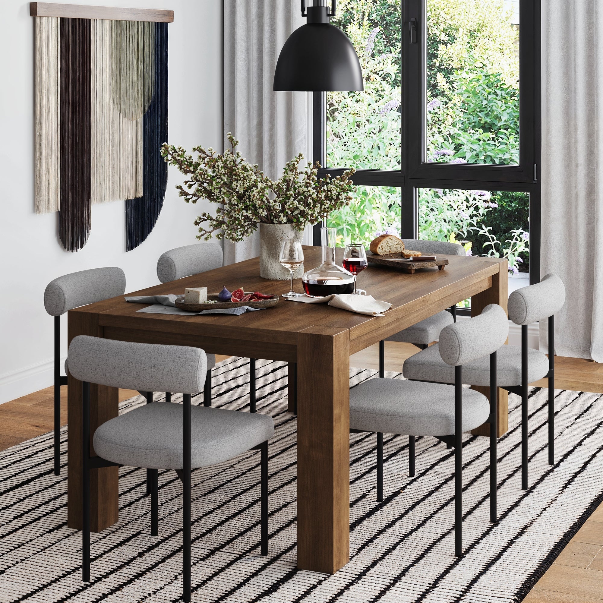 A modern dining room with a wooden table, six Nathan James Metal Modern Dining Chairs in gray, striped rug, black pendant light, and wall hanging. The table holds wine glasses, bread on a board, and flowers; large windows show greenery outside.