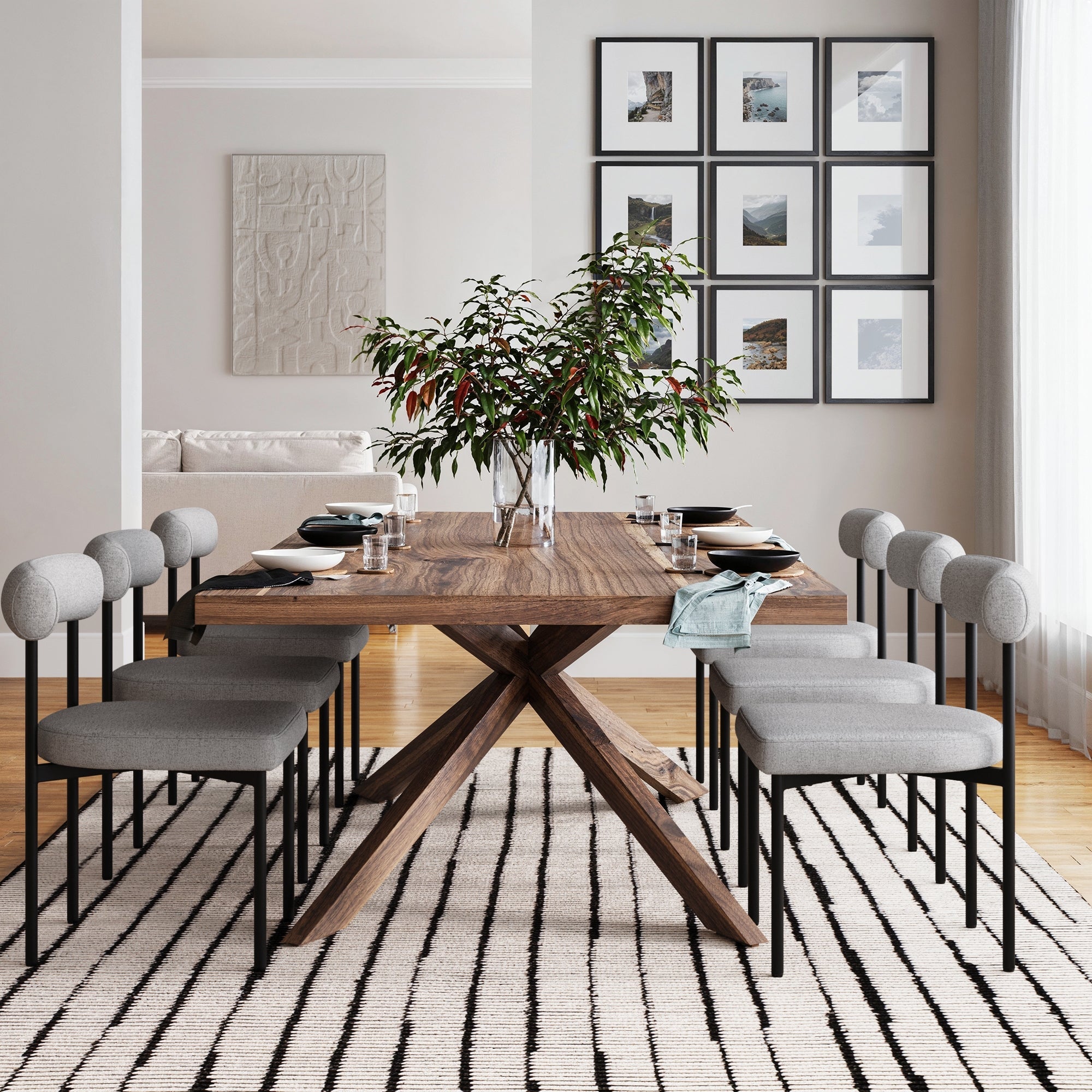 A modern dining room with a wooden table, six gray Nathan James Metal Modern Dining Chairs, a vase of leafy branches, striped rug, wall art, and large windows streaming in natural light.