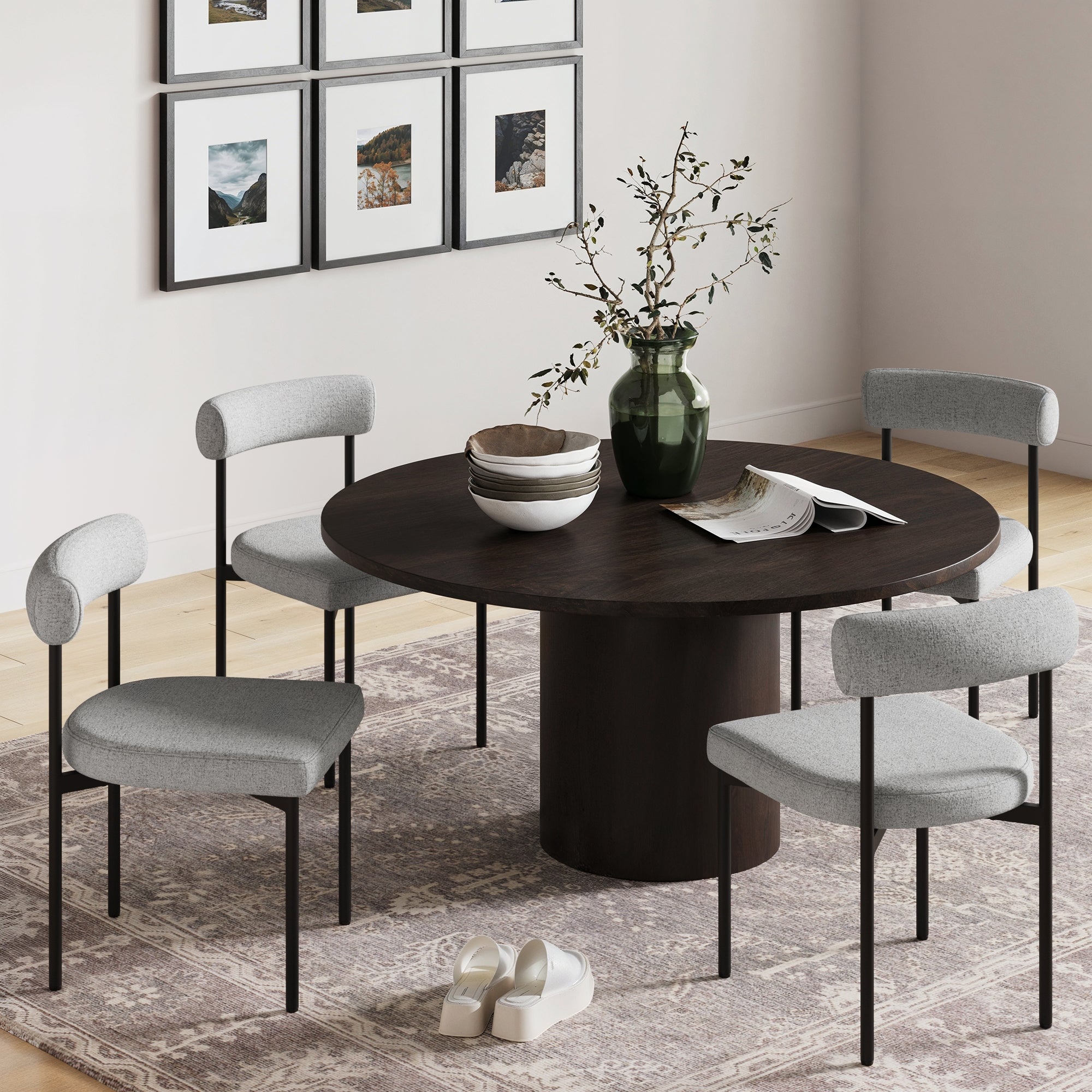 A round dark wood dining table with four Nathan James Metal Upholstered Modern Dining Chairs in gray sits on a patterned rug. On the table are a green vase with branches, bowls, and a magazine; shoes and framed photos are nearby.