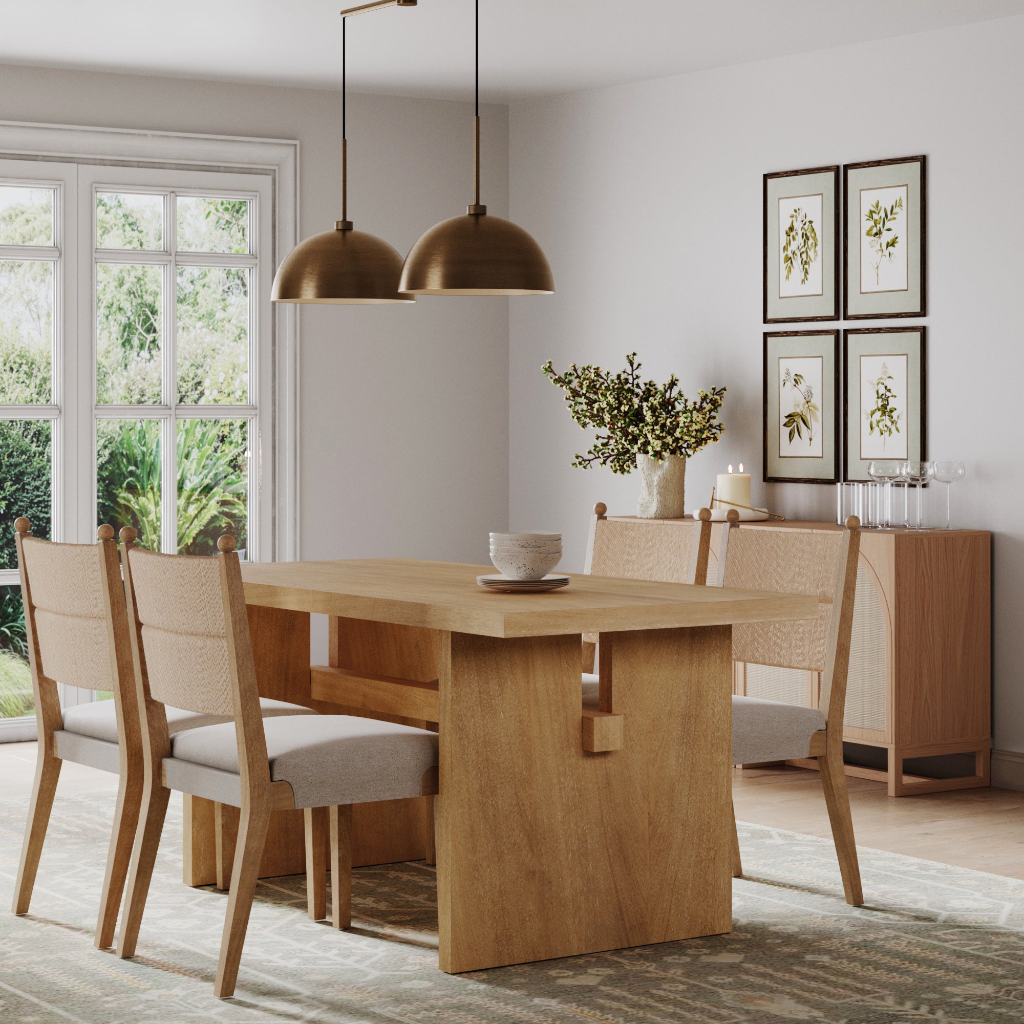 A modern dining room features a wooden table, a sideboard, brass pendant lights, botanical art, greenery in a vase, garden views through large windows, and four Nathan James Boho Natural Flax & Seagrass Dining Chairs.