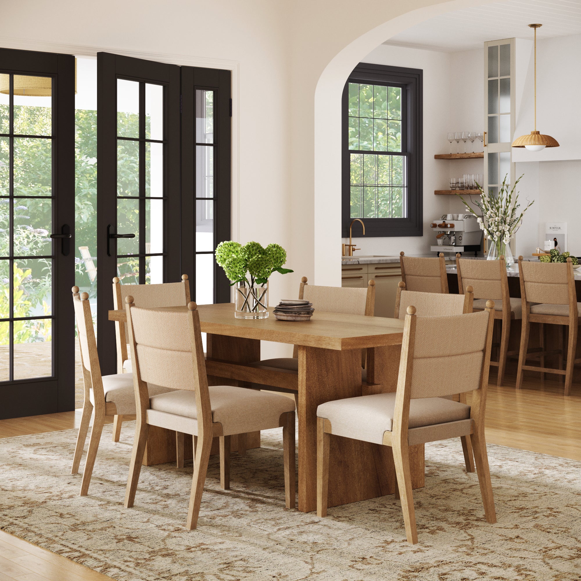 A modern dining room features a wooden table, a patterned rug, green flowers in a vase, and six Nathan James Boho Natural Flax & Seagrass Dining Chairs. Glass doors lead to the garden, with the kitchen visible beyond.