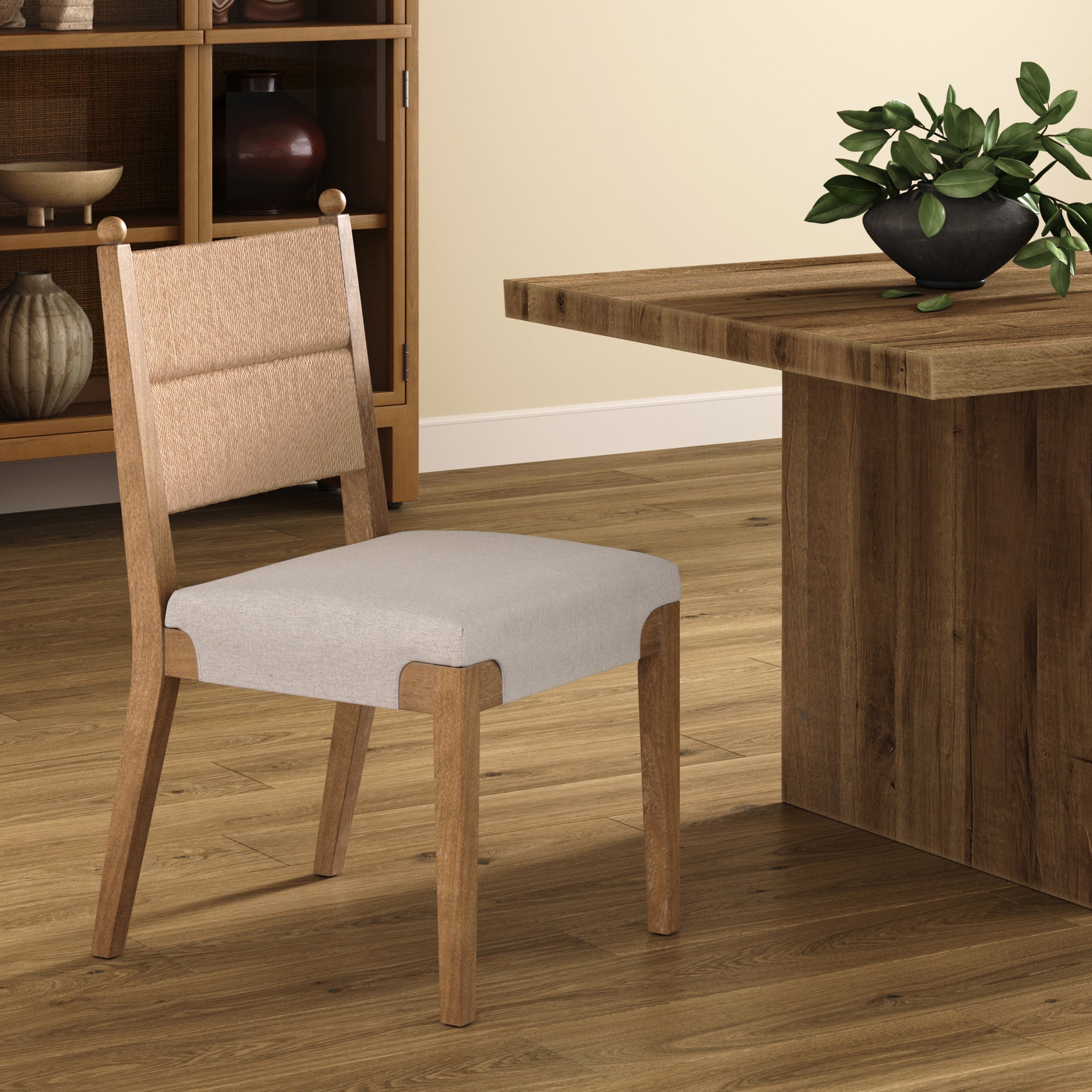 The Boho Natural Flax & Seagrass Dining Chair by Nathan James features a solid wood frame and cushioned beige seat, placed beside a wooden table on hardwood floors, with a potted plant and wooden cabinet in the background.
