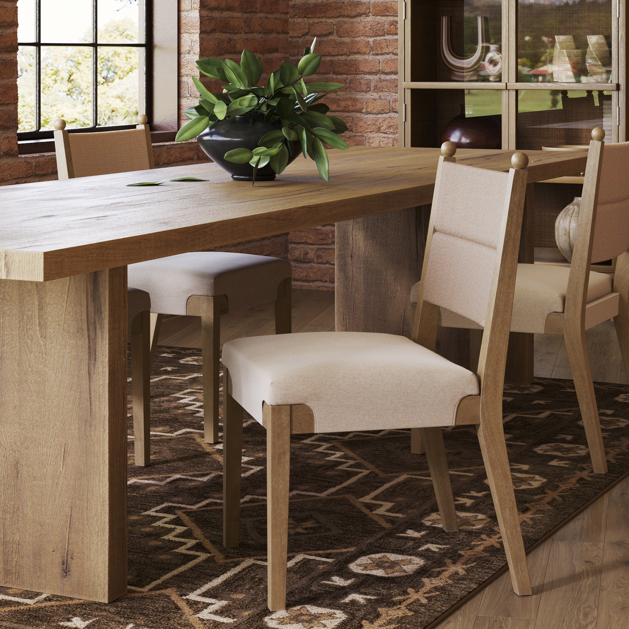 A wooden dining table with Nathan James Boho Natural Flax & Seagrass Dining Chairs sits on a patterned rug in a bright room. The chairs feature beige cushions and seagrass backs, while a black bowl with green leafy plants decorates the table.